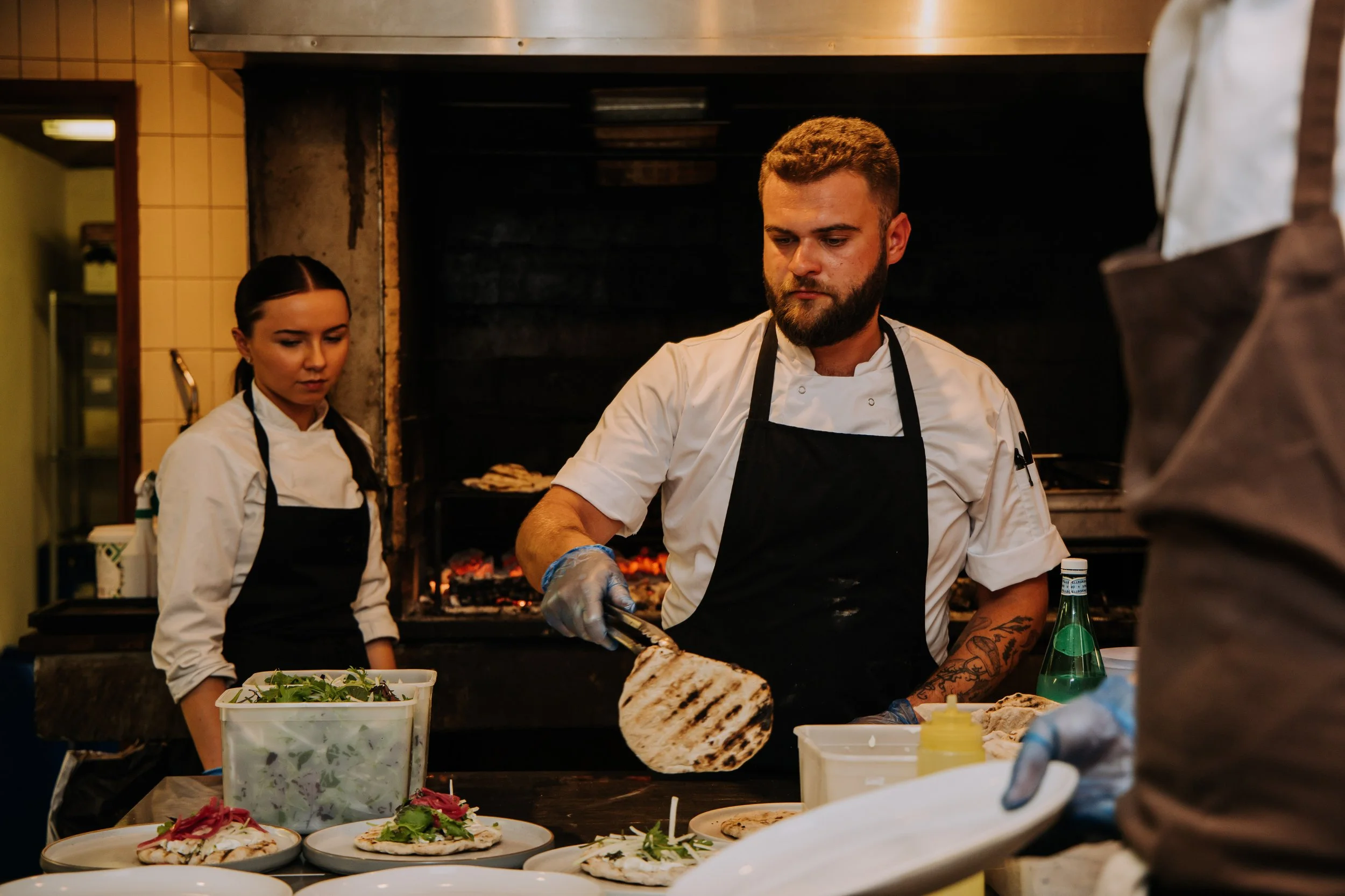 Chef cooking grilled chicken in restaurant kitchen with two women preparing salads in the background.