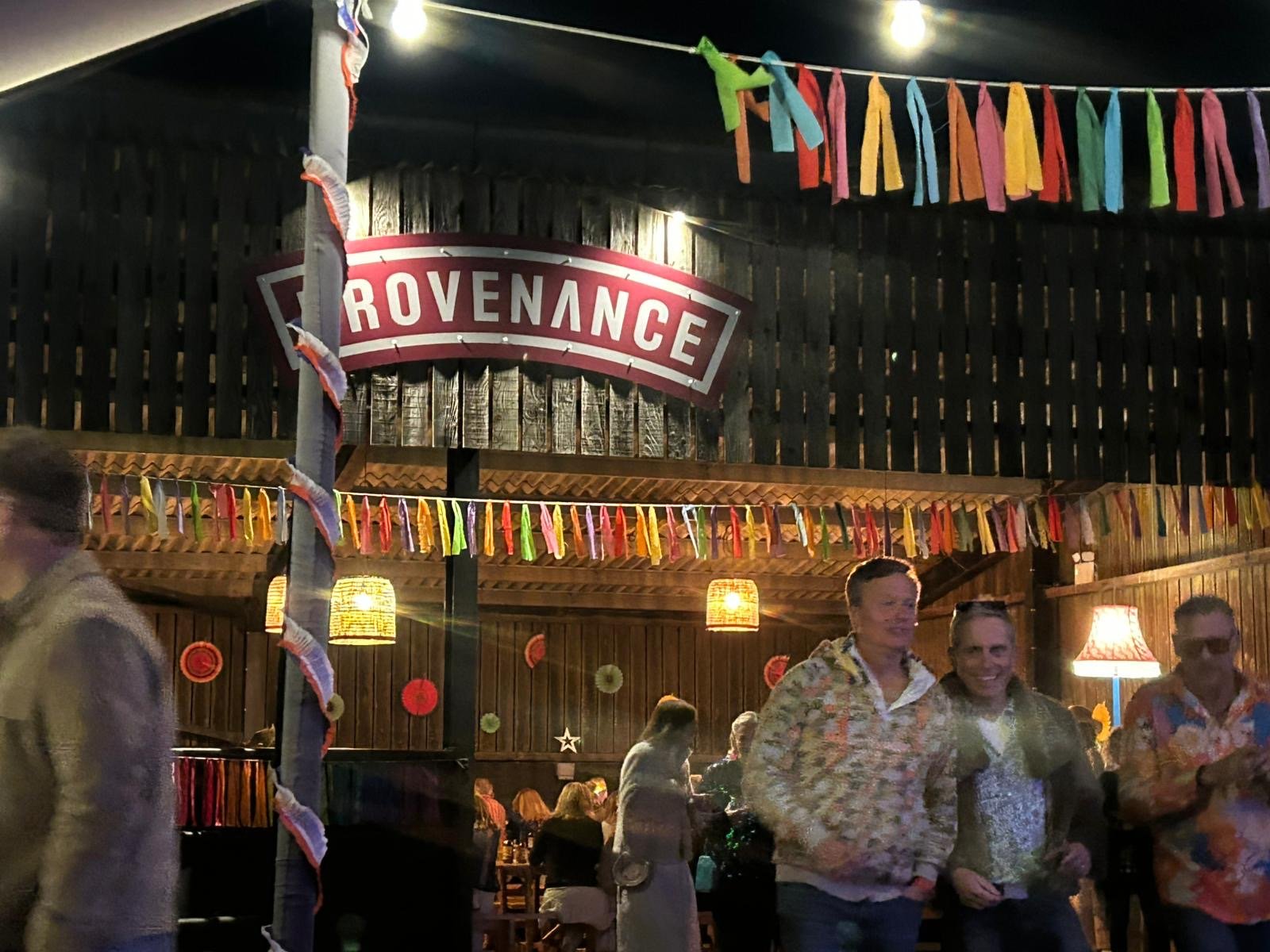 A social gathering at a venue with colorful decorations, hanging paper fans, and a large sign that reads 'Provenance'. People are standing and conversing, some dressed in casual shirt patterns.