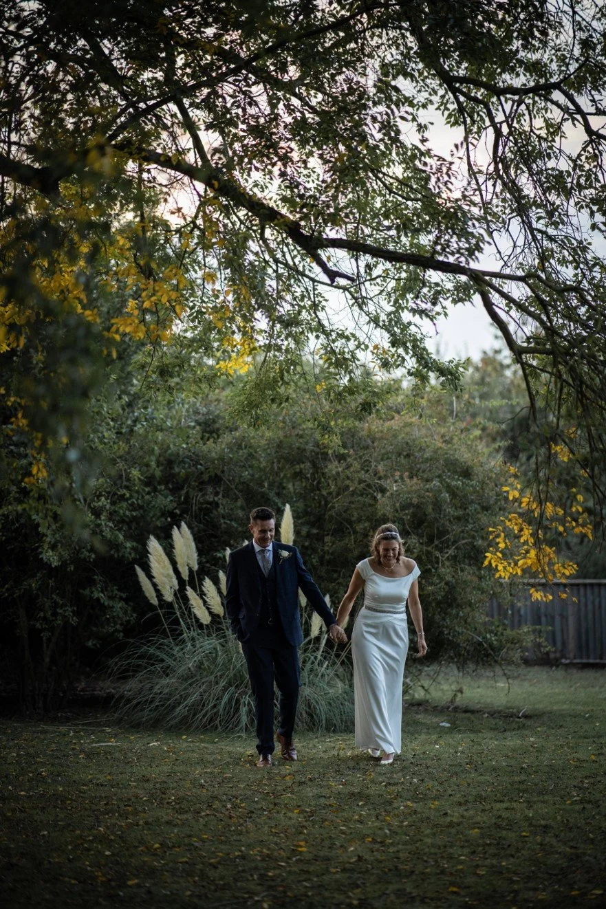 A bride and groom holding hands walking across a grassy field outdoors with trees and foliage in the background.