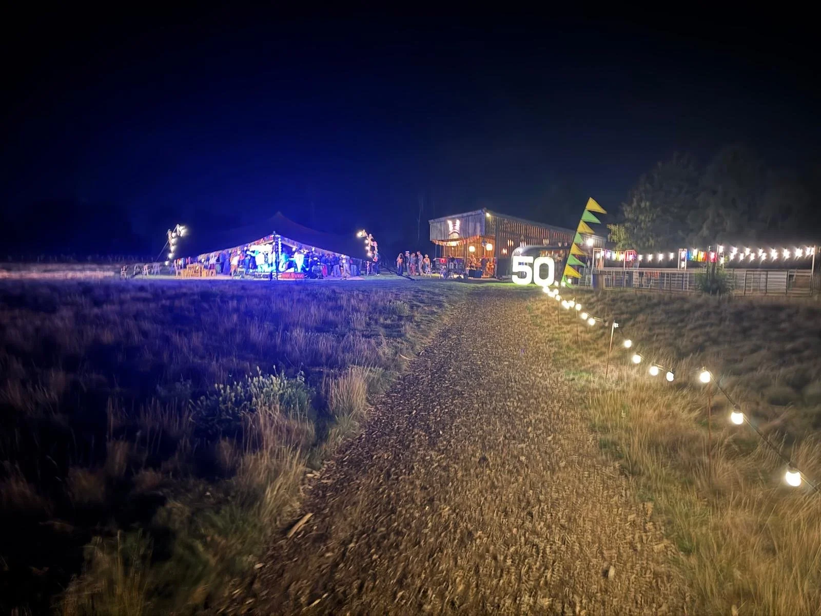 Night scene of a celebration with tents and string lights, a large illuminated '50' sign, and a gravel path leading to the event area.