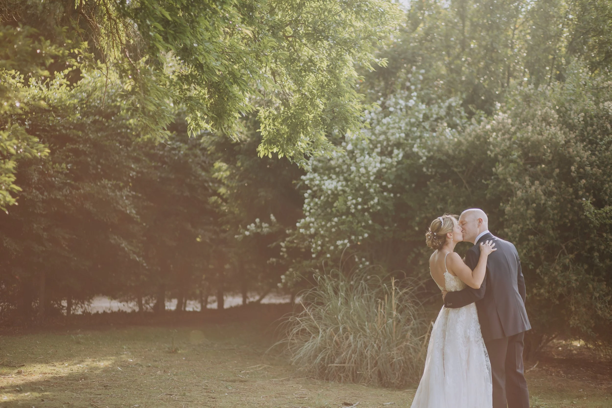 A bride and groom sharing a kiss in a lush outdoor setting with trees and greenery, during sunset or golden hour.