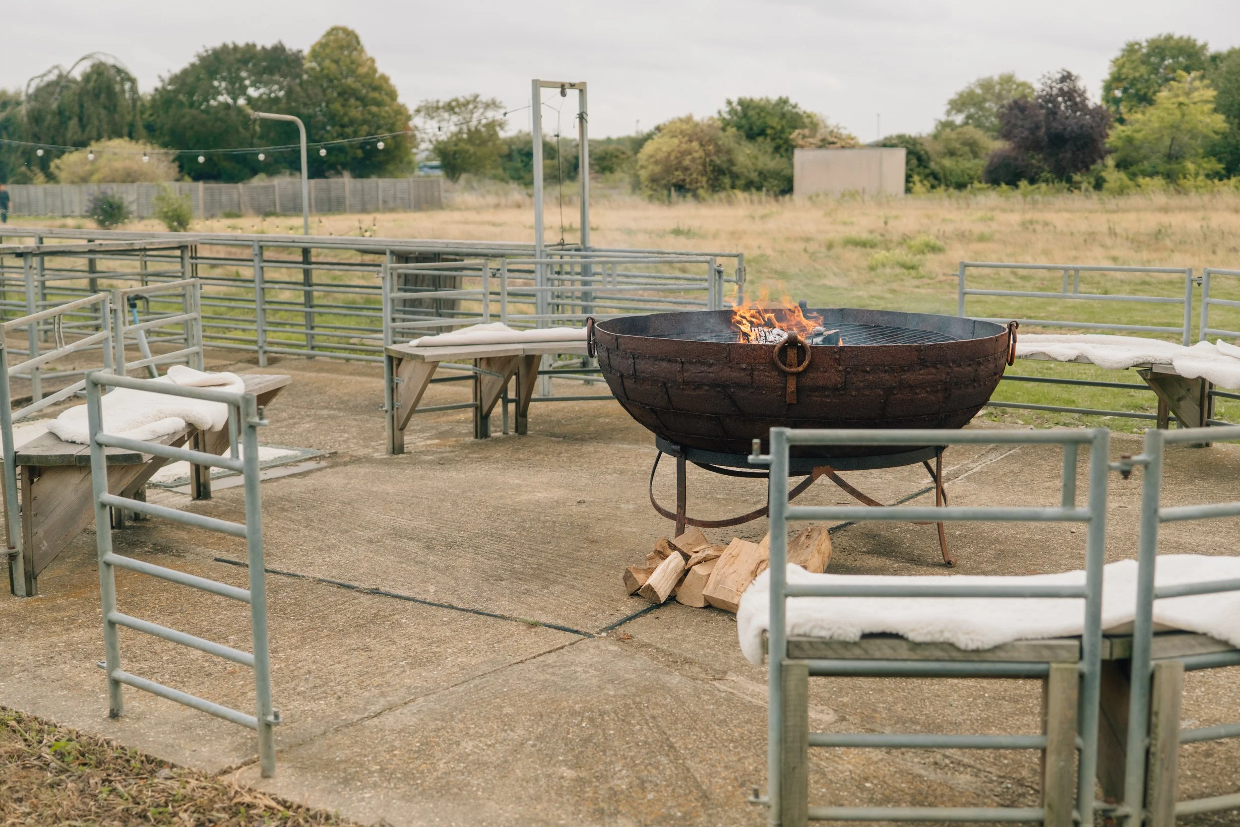 Outdoor seating area with benches and a fire pit in the center with a small fire burning, surrounded by a metal fence, with trees and open field in the background.
