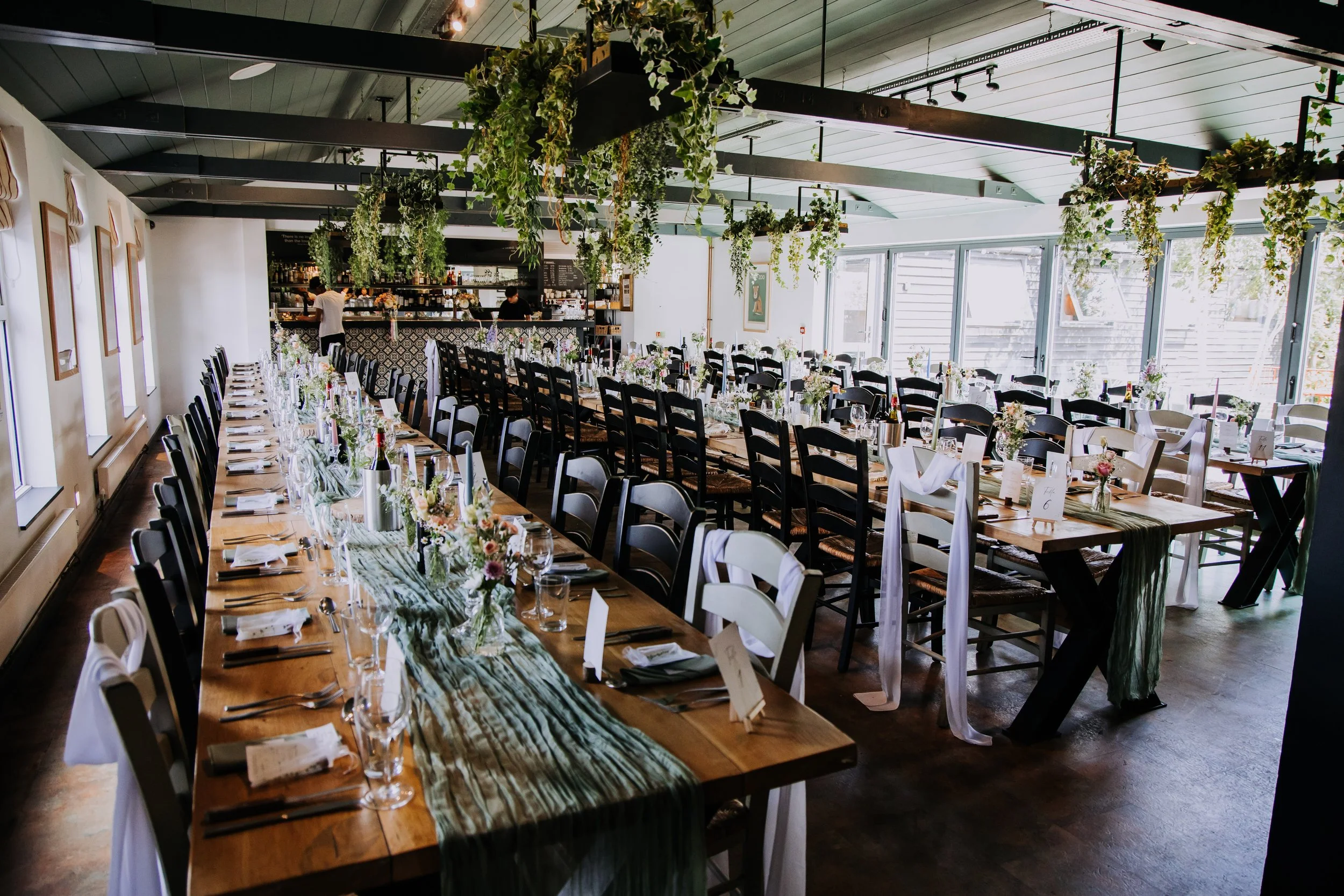 A decorated dining area with long tables and chairs, floral centerpieces, hanging greenery, and sunlight coming through large windows, set up for a celebration or event.