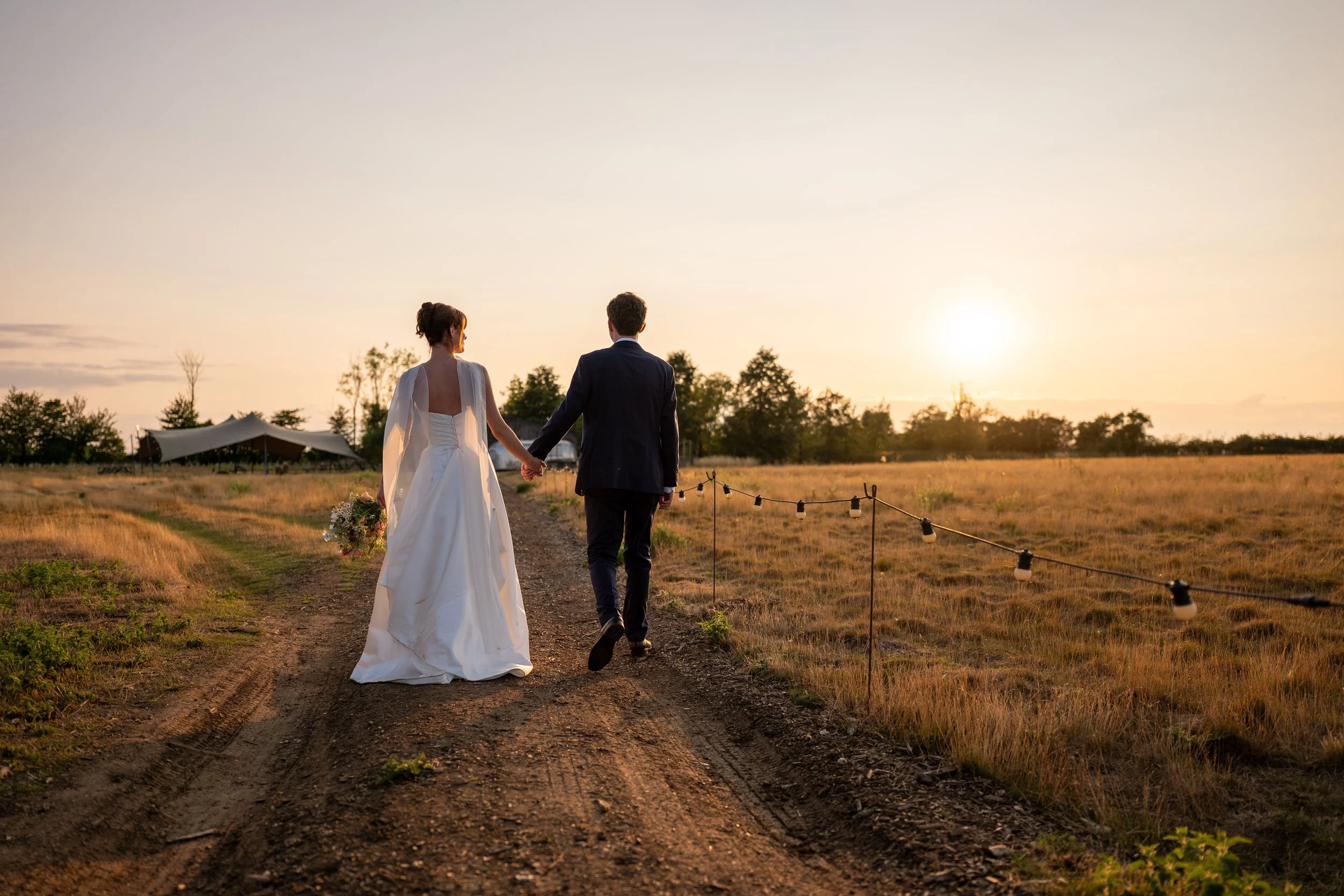 Bride and groom holding hands walking on a dirt path at sunset in a field.