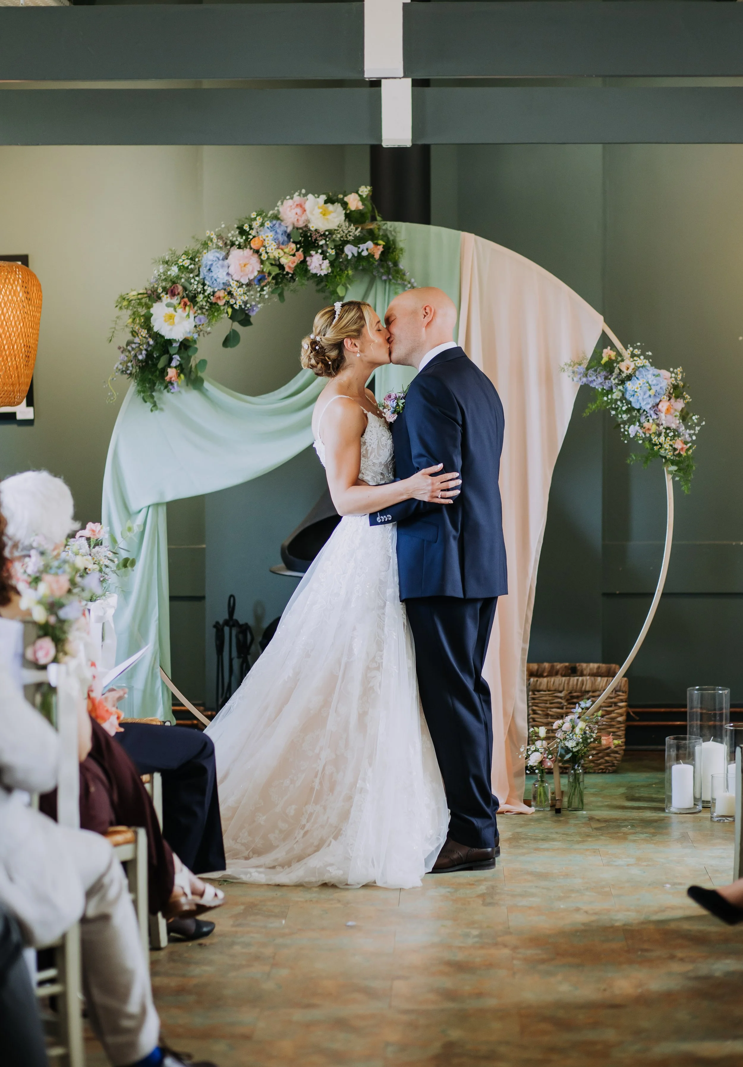 Bride and groom sharing a kiss during their wedding ceremony, standing beneath a floral arch with light green and beige fabric, with guests watching.
