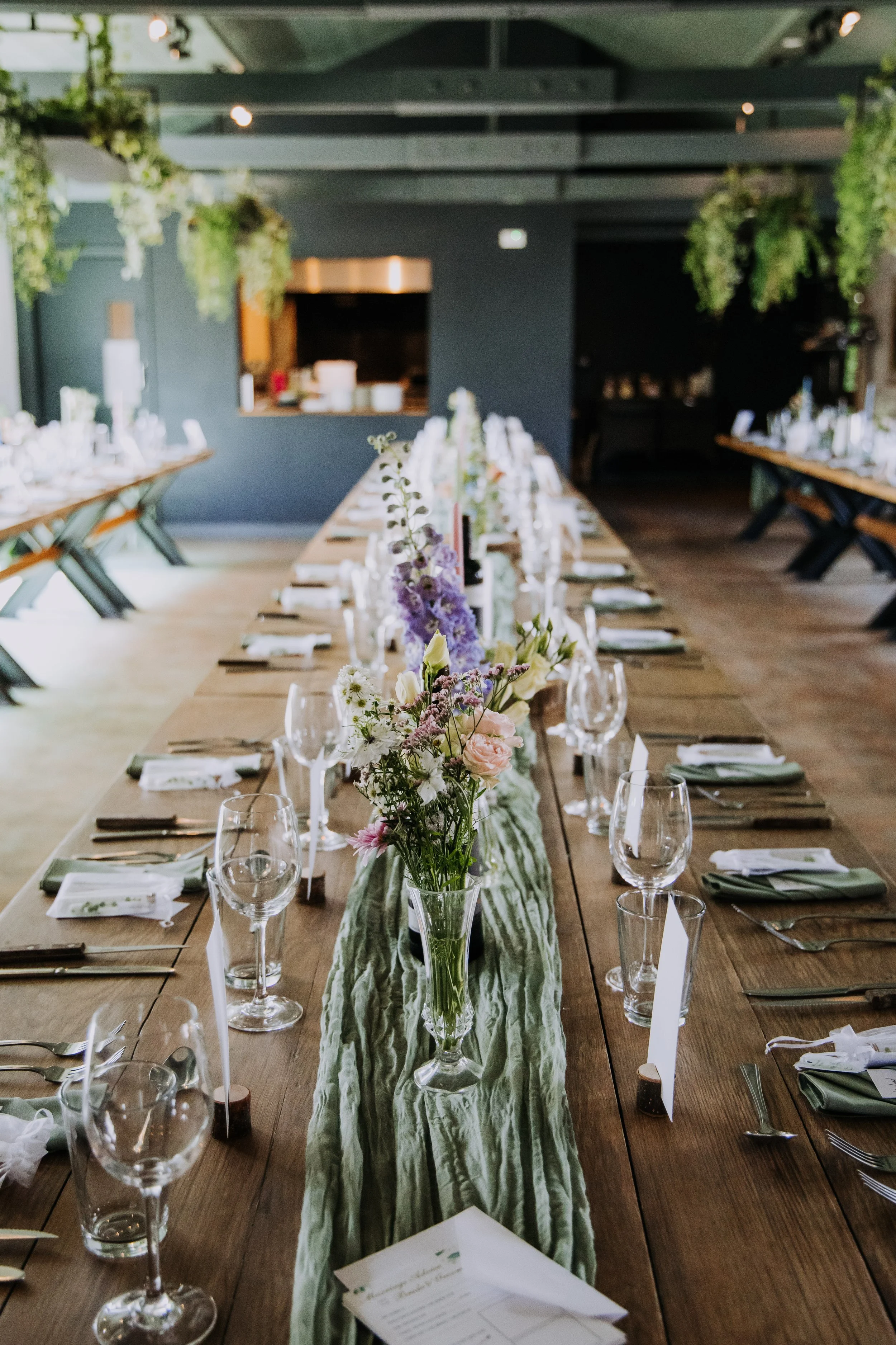 Long wooden dining table set for a formal event, decorated with a green fabric runner and a flower arrangement in the center. The table is arranged with wine glasses, plates, napkins, and silverware, in a room with dark walls and hanging greenery.