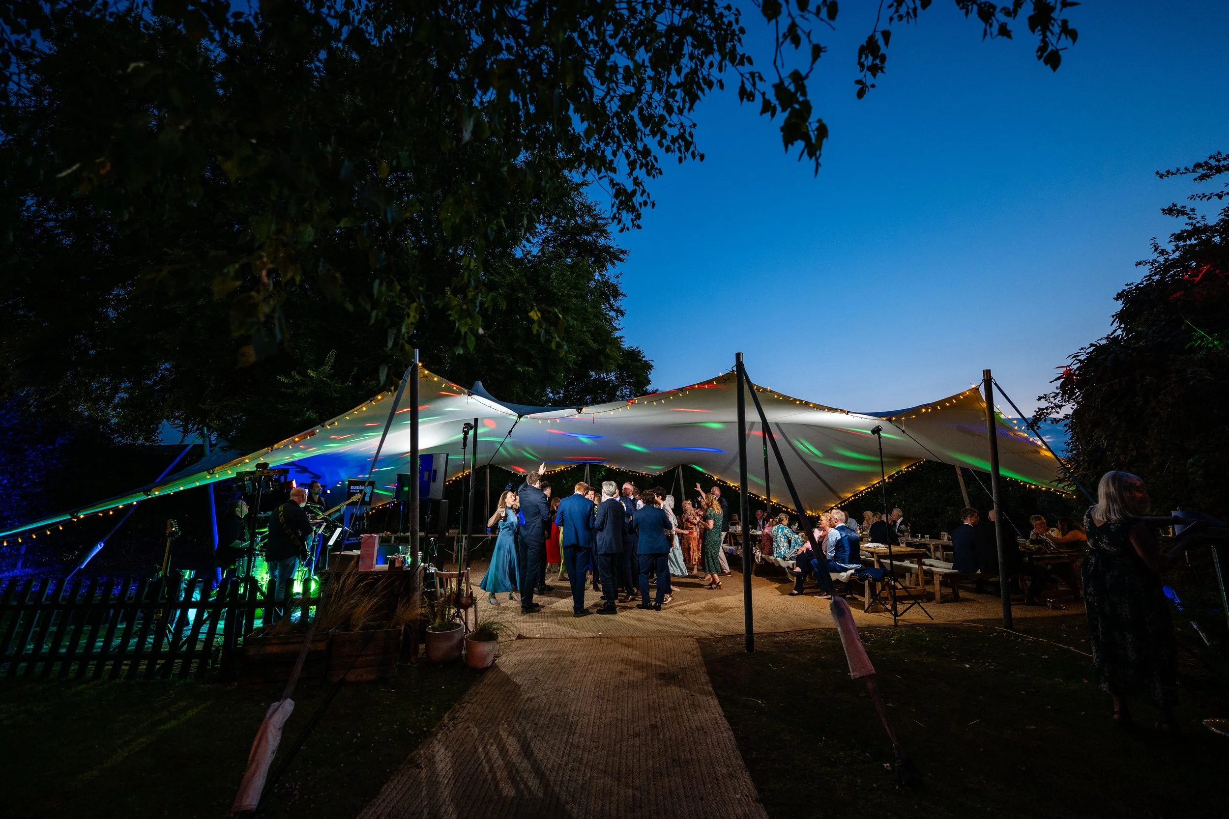 People dancing and socializing under a large illuminated canopy at an outdoor evening event, with trees and a blue sky in the background.