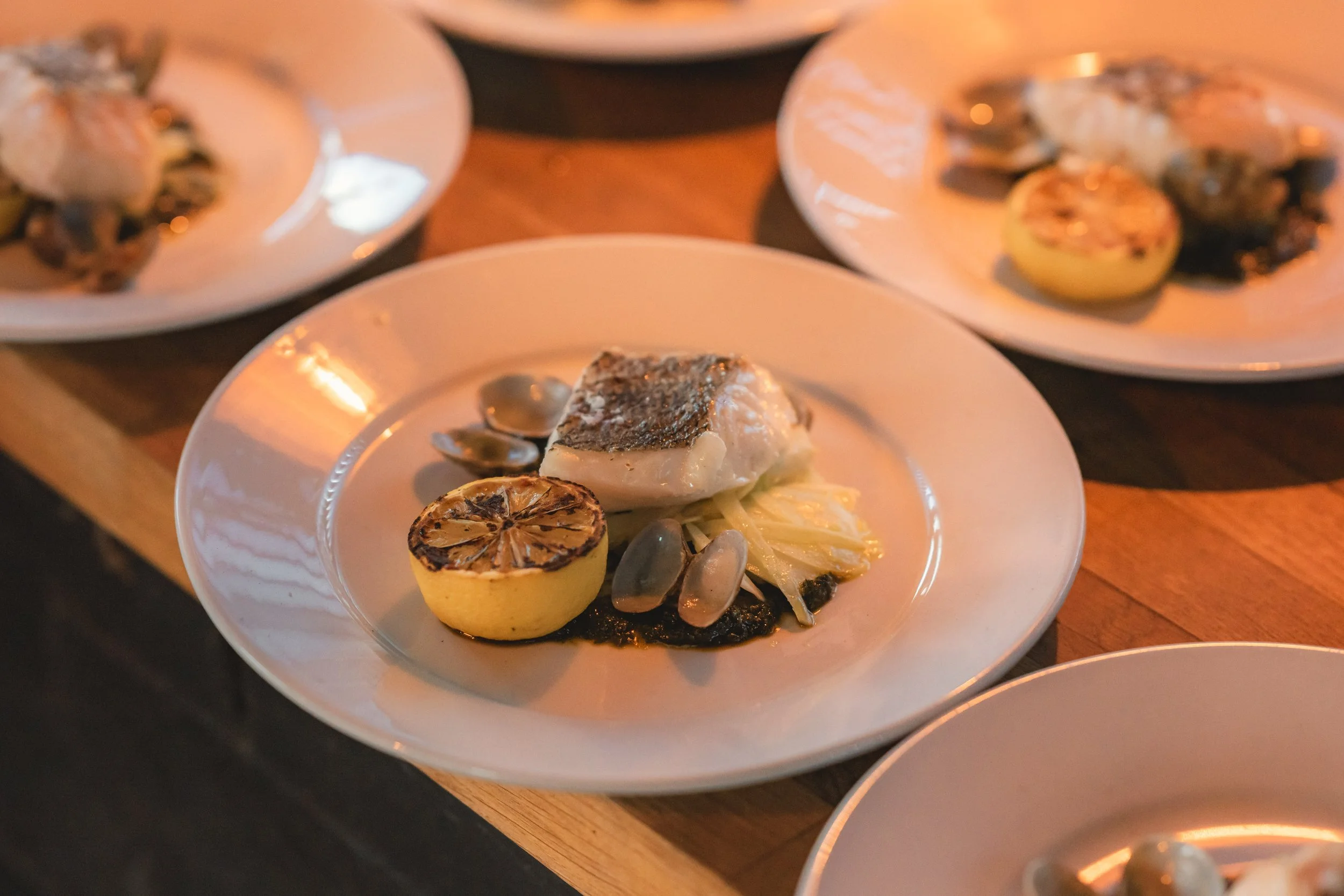A white plate with a cooked fish fillet, grilled lemon half, roasted garlic, and some vegetables on a wooden table.