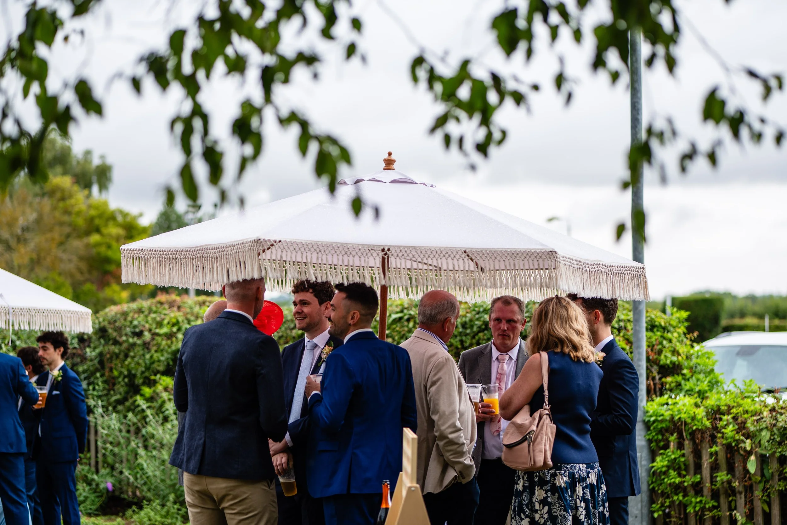 Group of people socializing outdoors under umbrellas, some holding drinks, with greenery and sky in background.