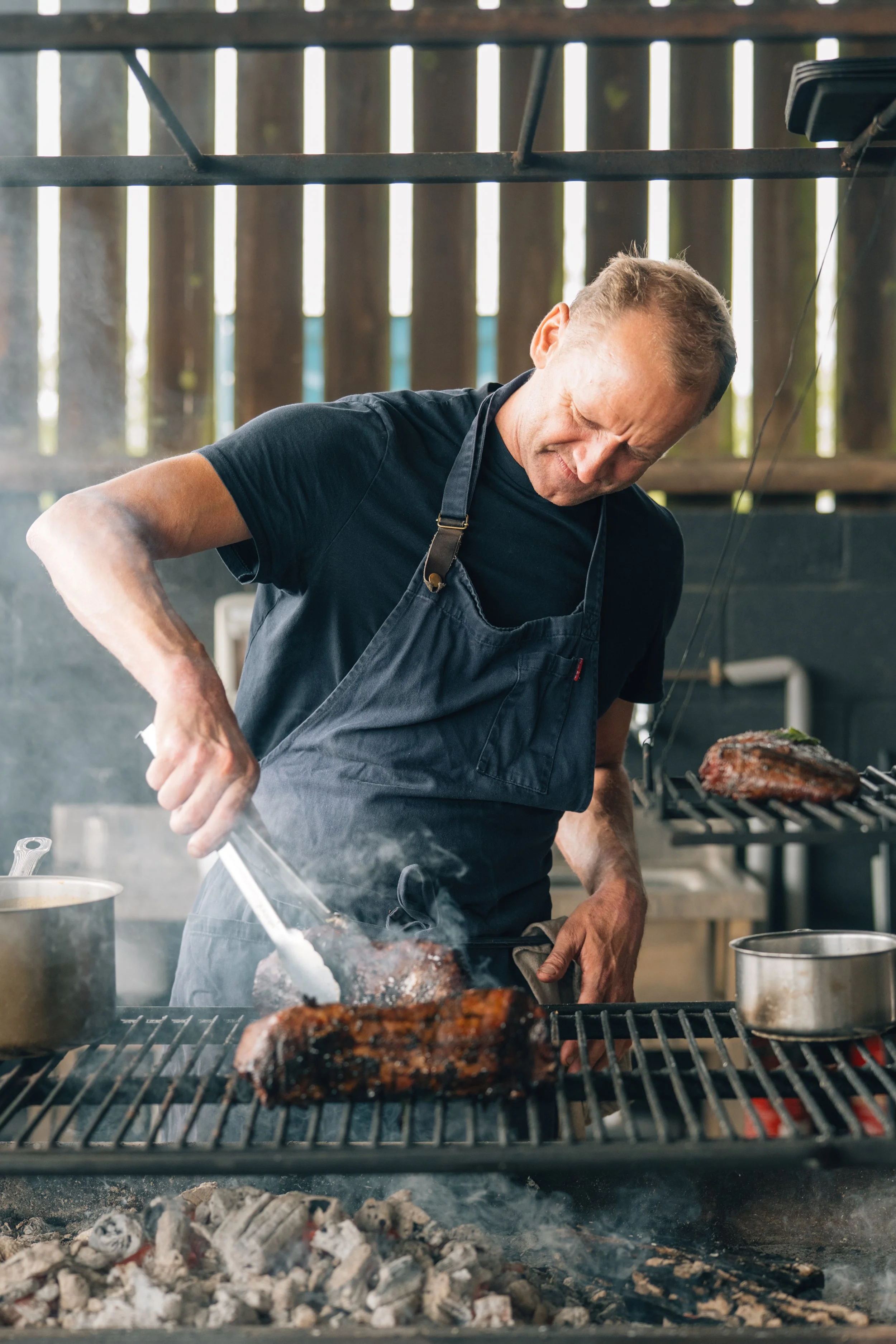 Man grilling meat on outdoor barbecue grill, wearing black apron, using tongs to turn meat, with wood fence in background.
