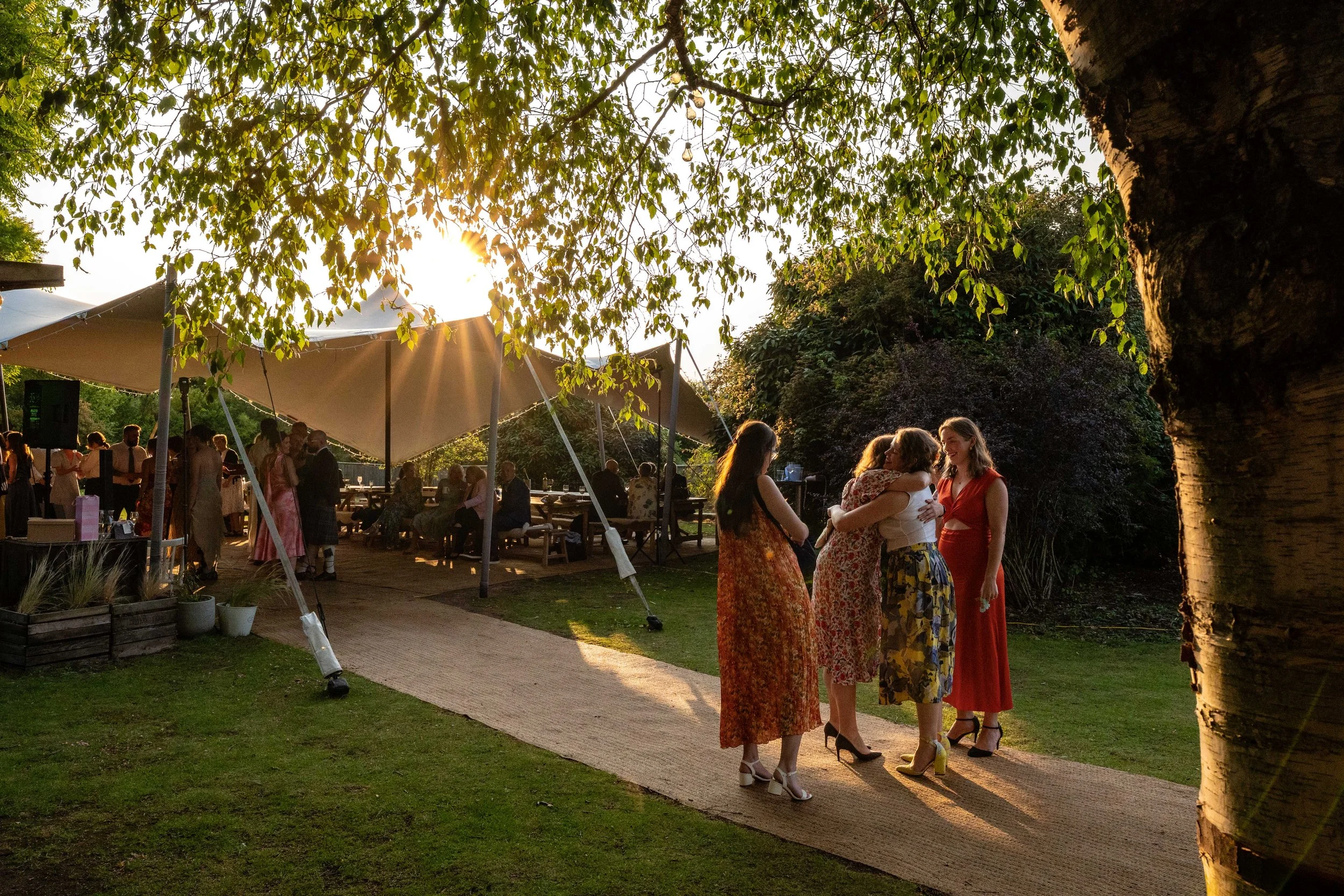People dancing and chatting at an outdoor event during sunset, with a large white tent and trees in the background.