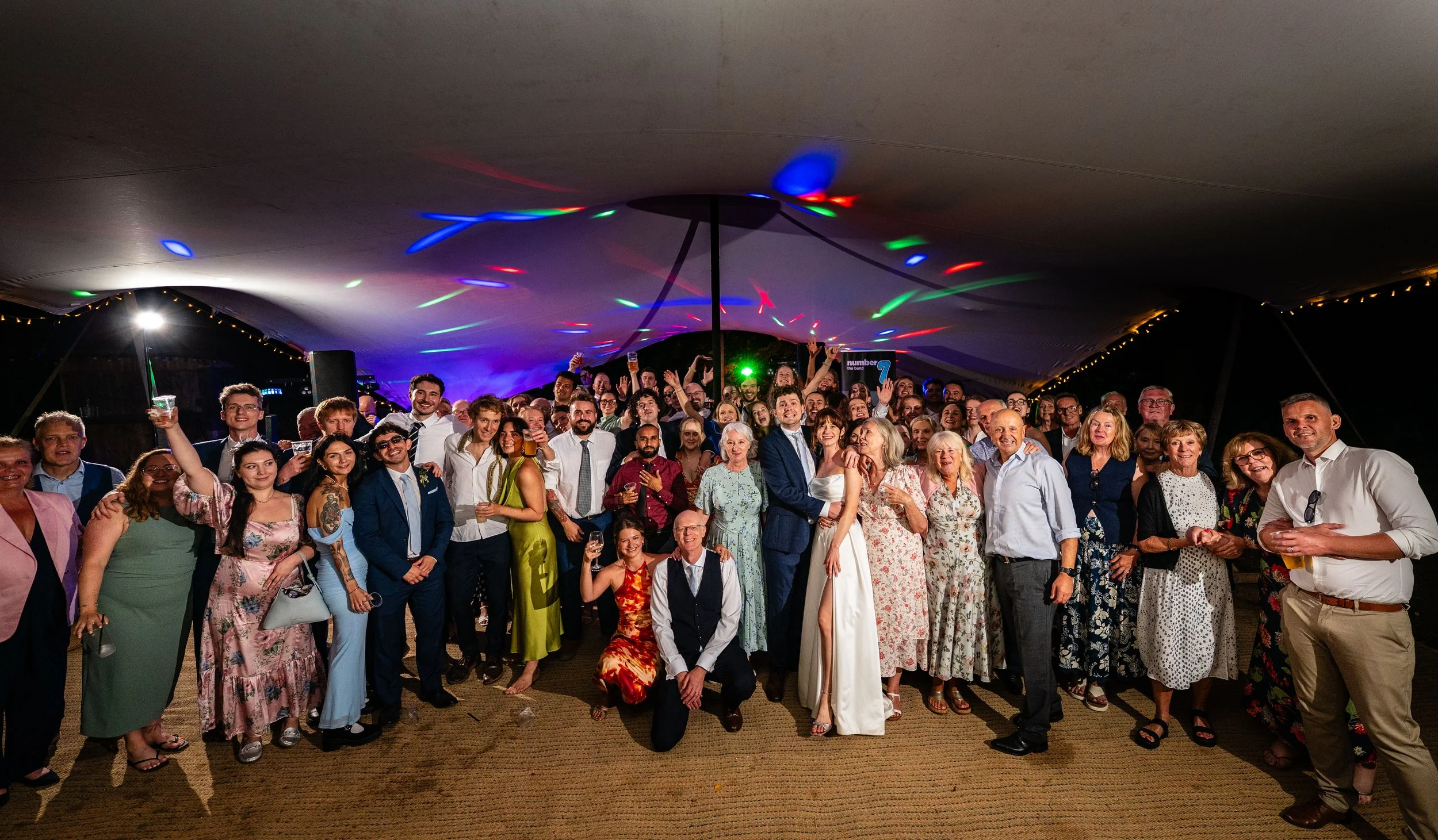 A large group of people posing together at a celebration under a decorated tent with colorful lights.