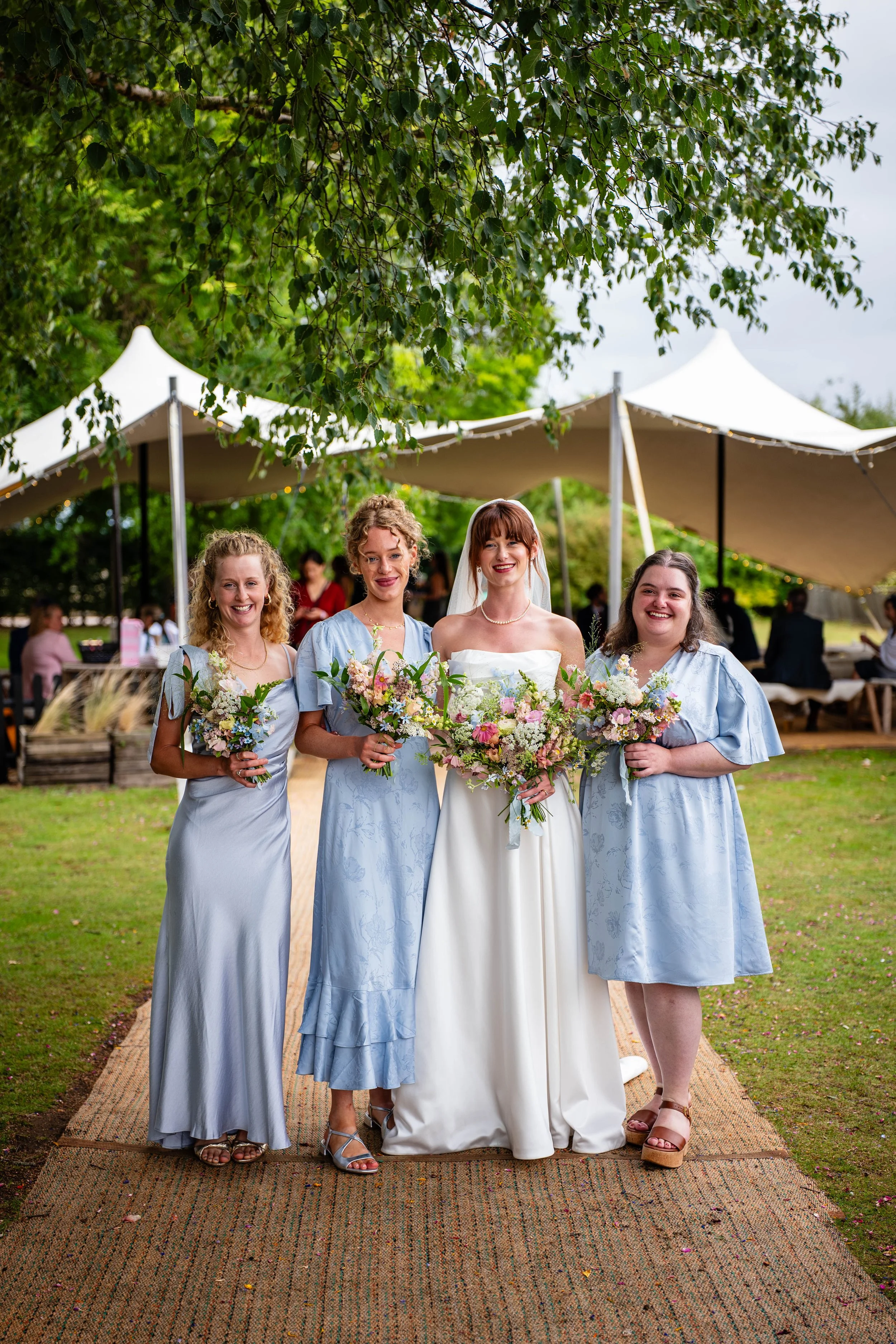 A bride and three bridesmaids smiling and holding bouquets at an outdoor wedding ceremony under a large white tent.