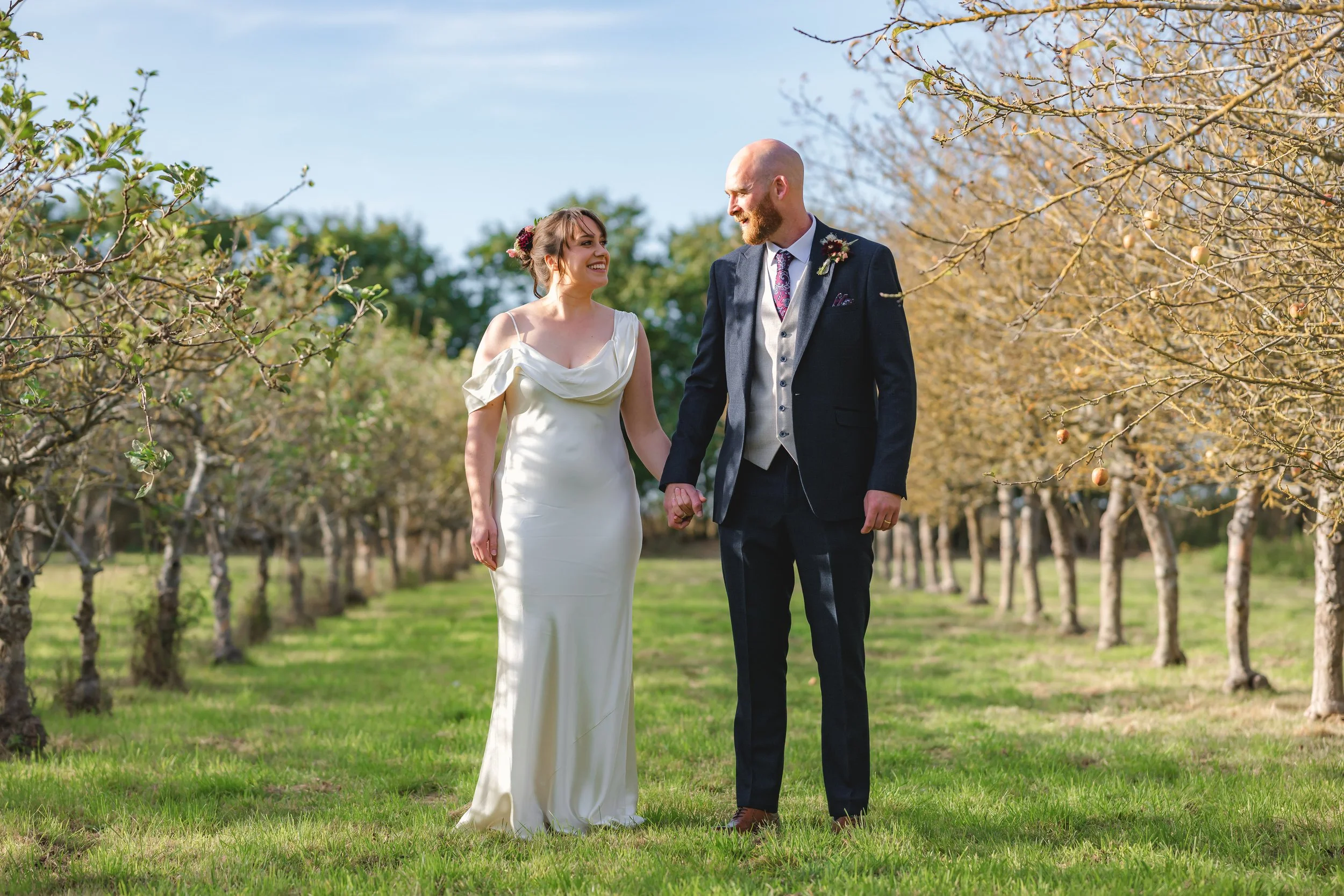 A bride and groom holding hands and smiling at each other in an orchard with leafless trees and green grass during daytime.