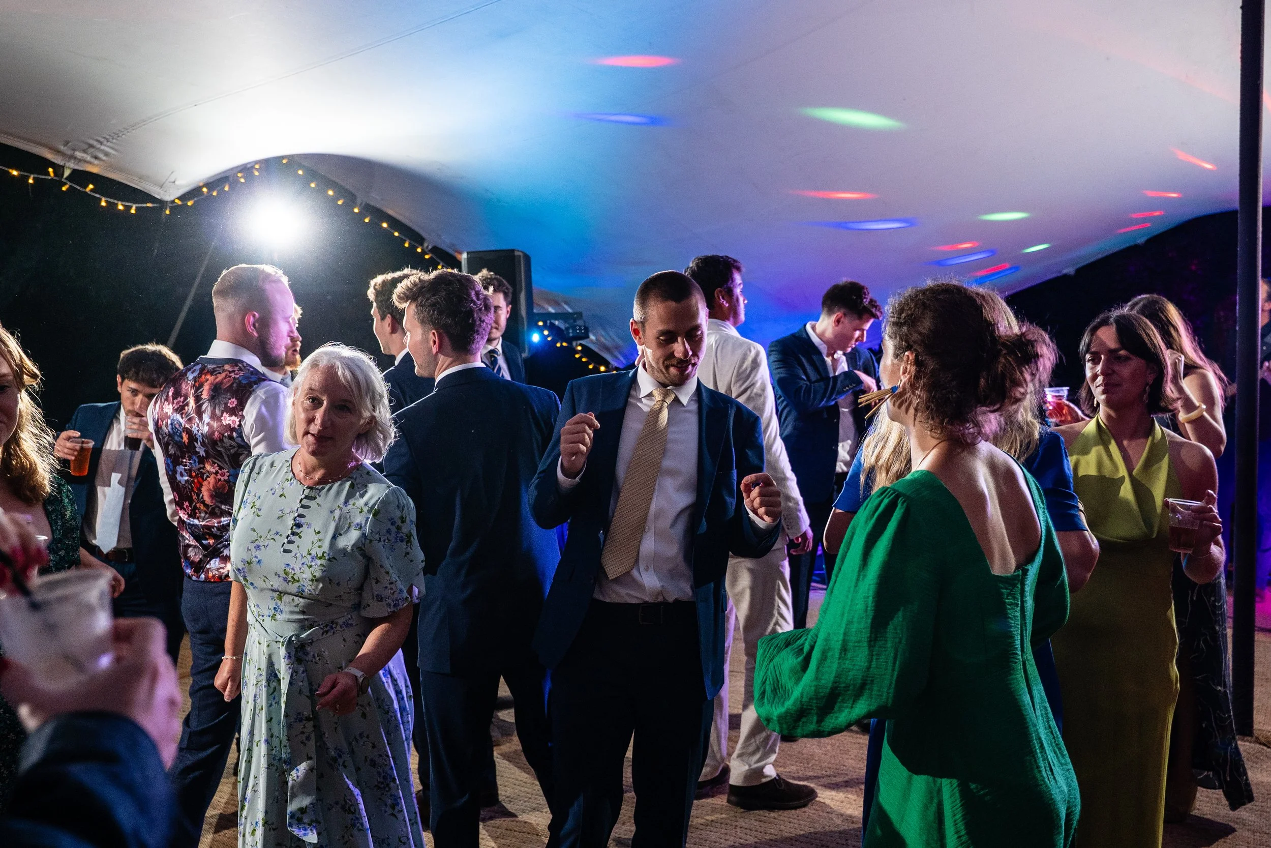 People dancing and socializing at an indoor party or reception under colorful lighting with a curved tent ceiling.