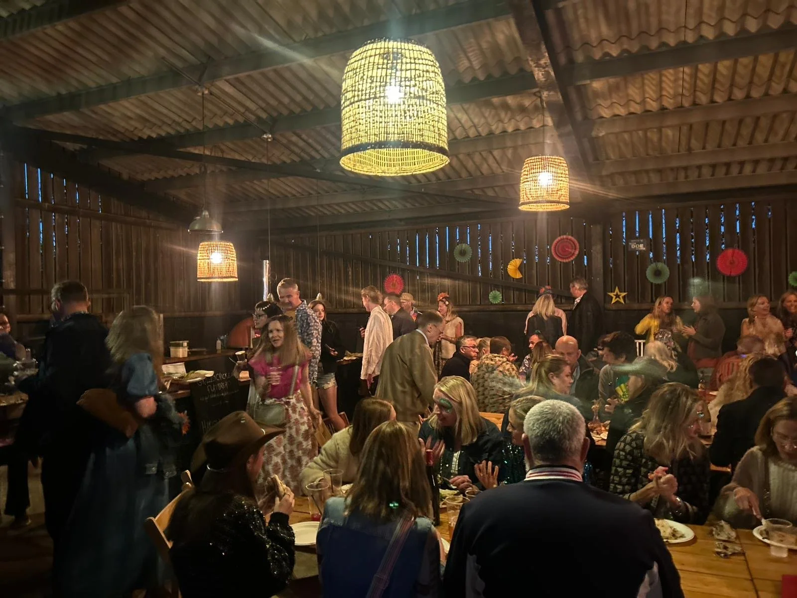People at a festive indoor gathering with wooden walls and ceiling, decorated with colorful paper lanterns and star ornaments, illuminated by warm hanging lights.