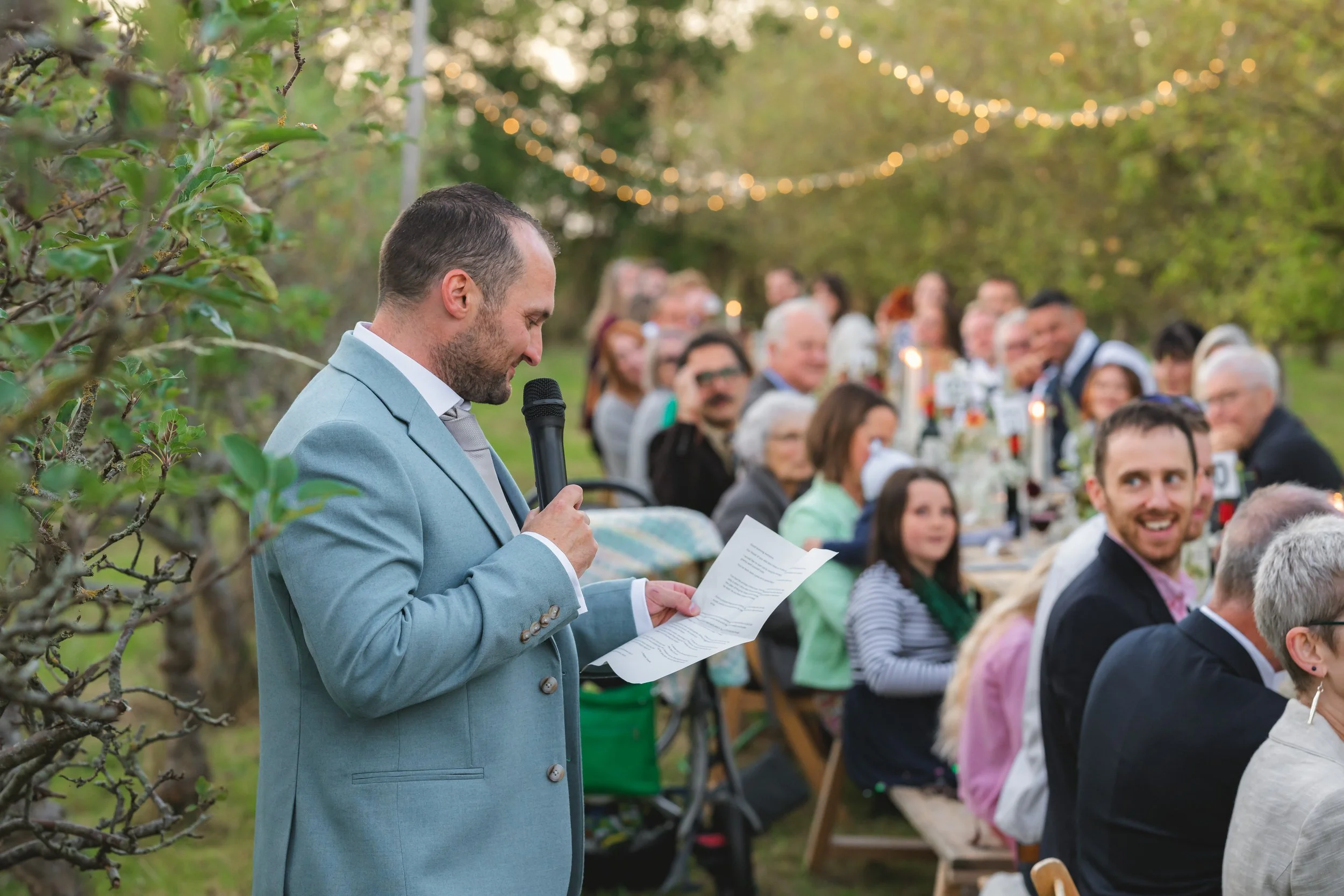 Man in light blue suit giving speech at an outdoor event with guests seated at long tables decorated with candlelights and flowers, surrounded by trees.