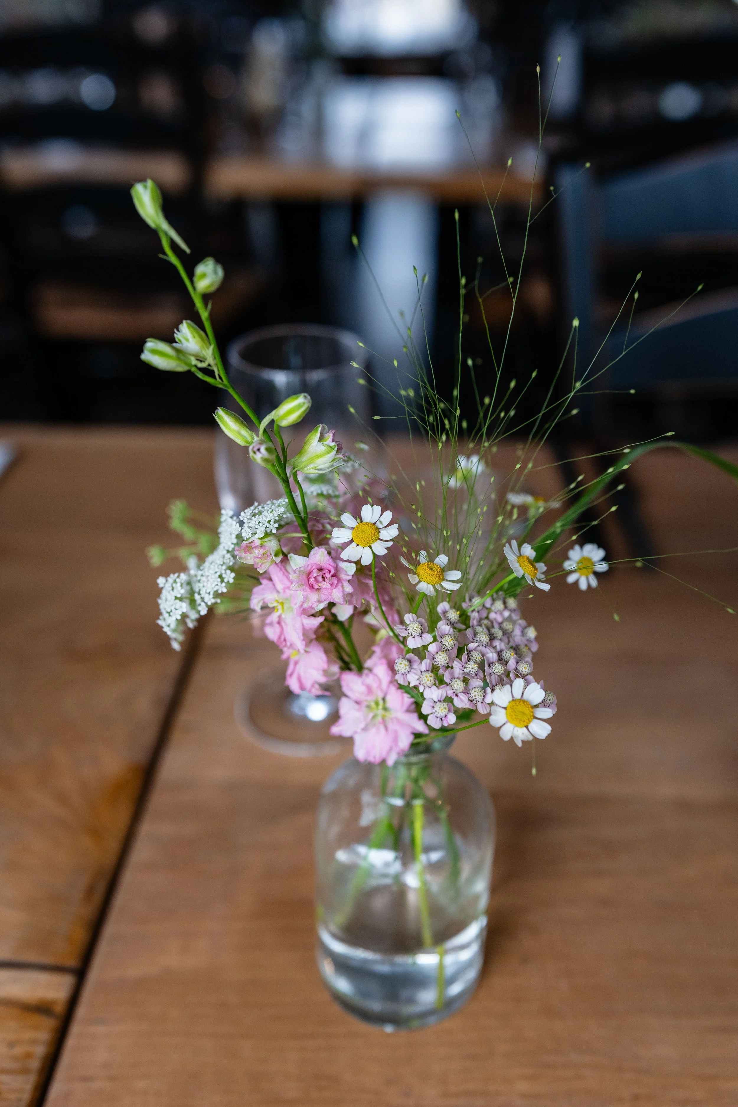 A small glass vase with mixed pink and white wildflowers, including daisies, on a wooden table.