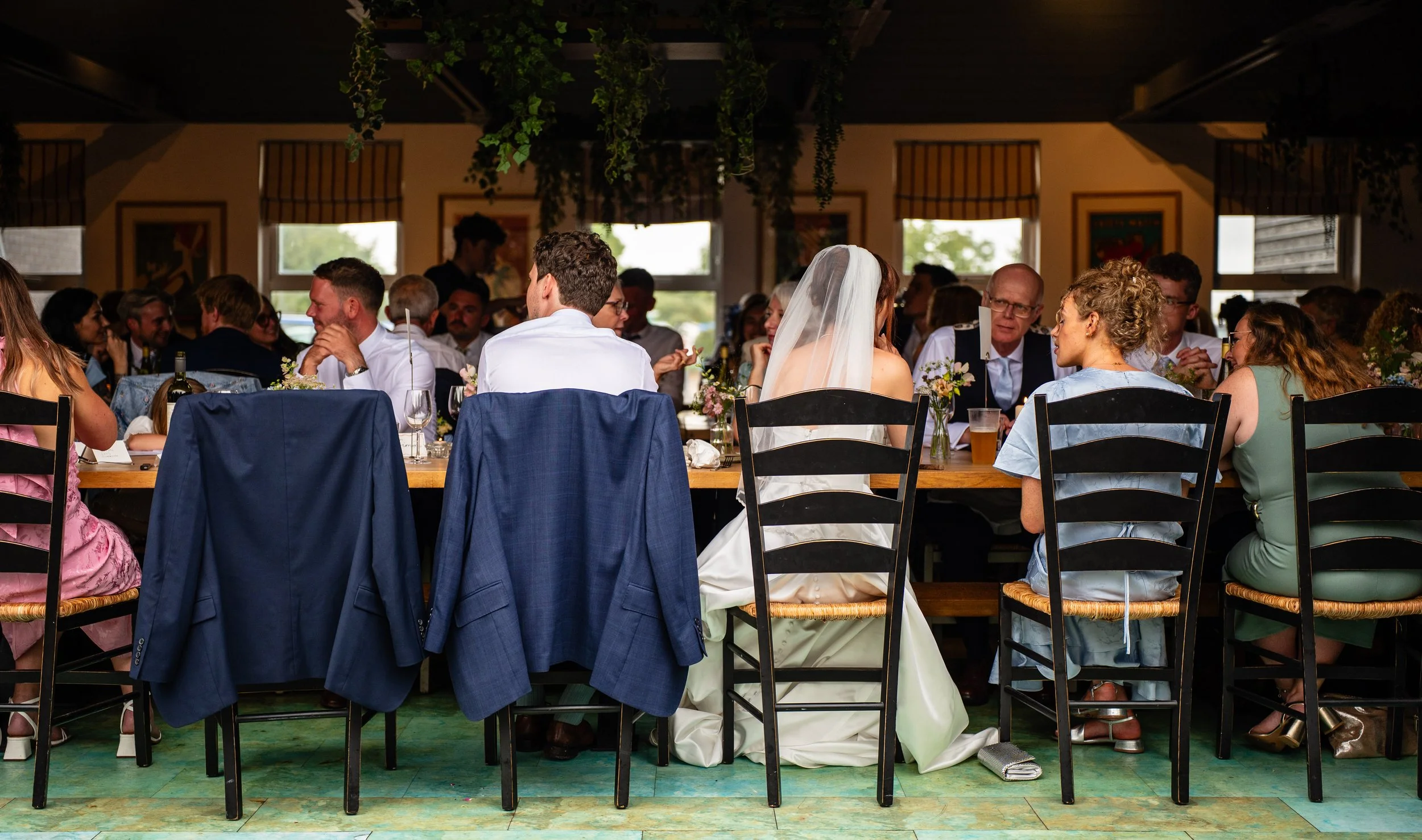 Wedding reception with guests seated at a long table, including the bride in a white dress with veil, and people engaged in conversation, in a decorated indoor venue.