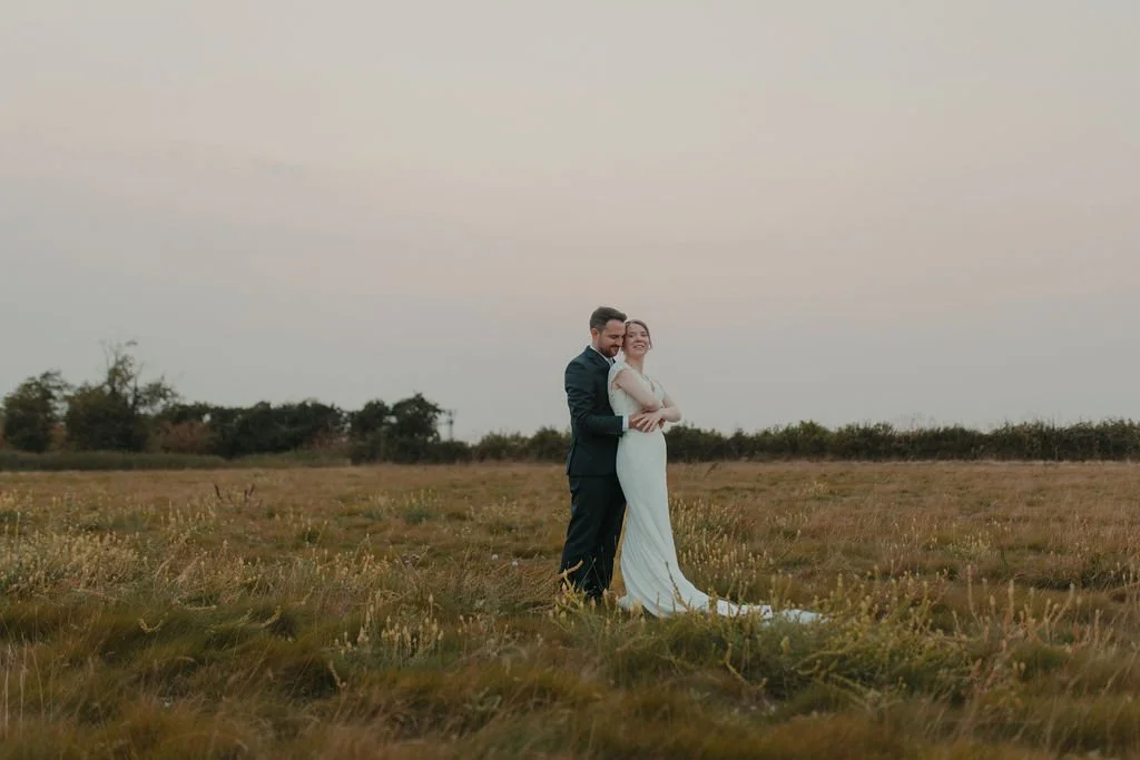 A bride and groom standing together in an open grassy field during sunset, smiling and embracing.