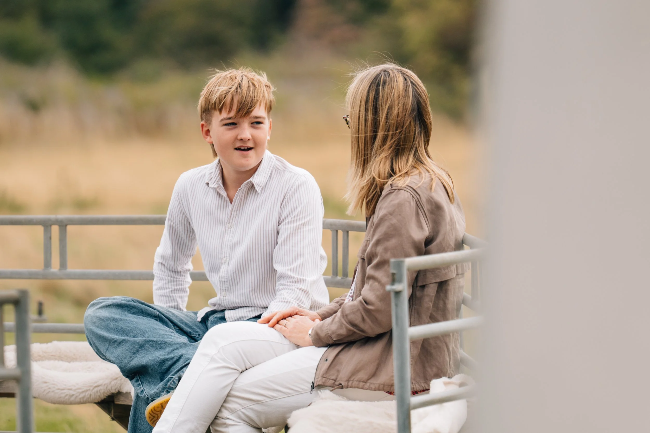 A young boy and woman sitting outdoors on a bench, engaged in conversation, with a blurred natural background.