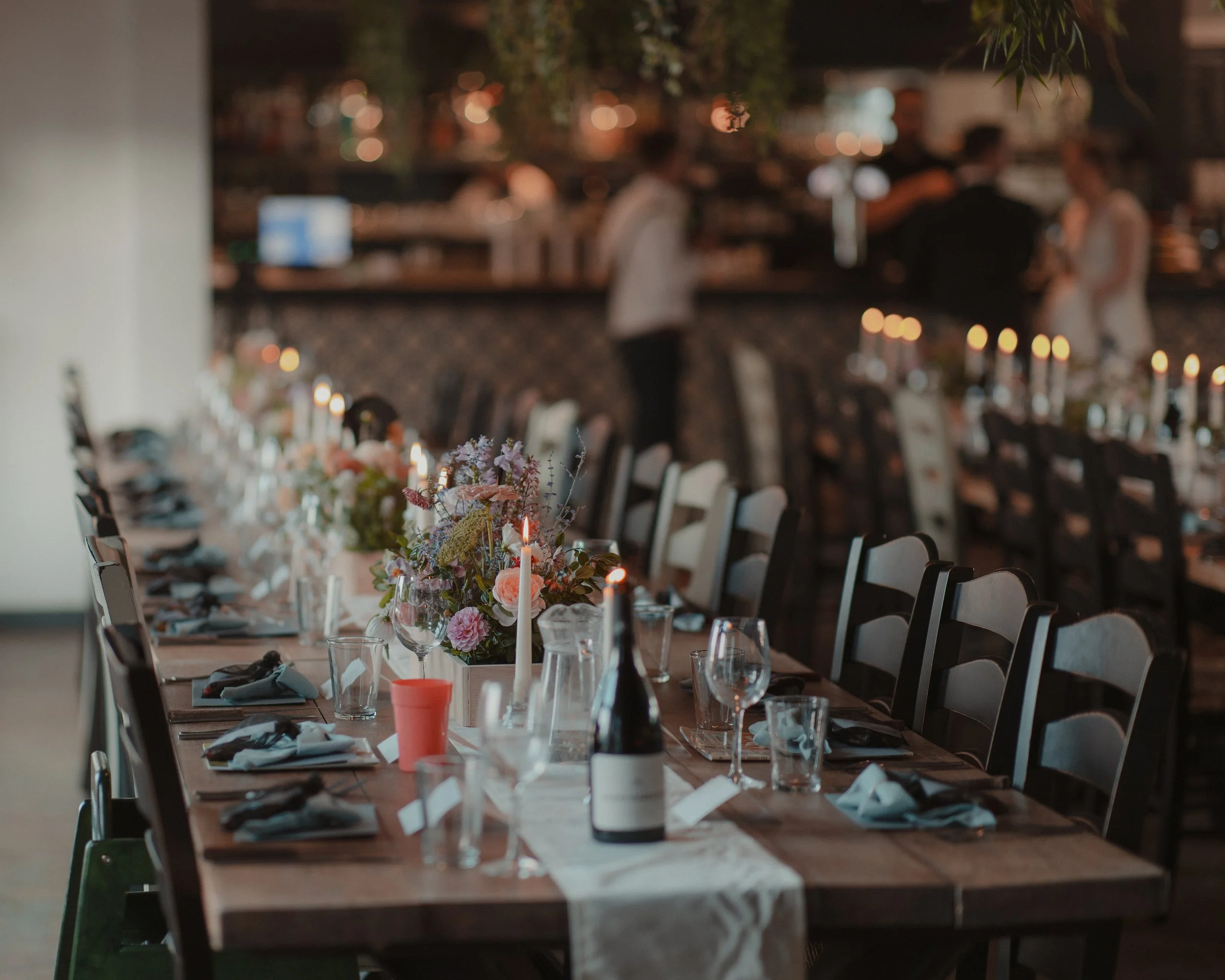 A long dining table decorated with floral centerpieces, candles, wine bottles, glasses, and place settings in a warmly lit venue with people in the background.