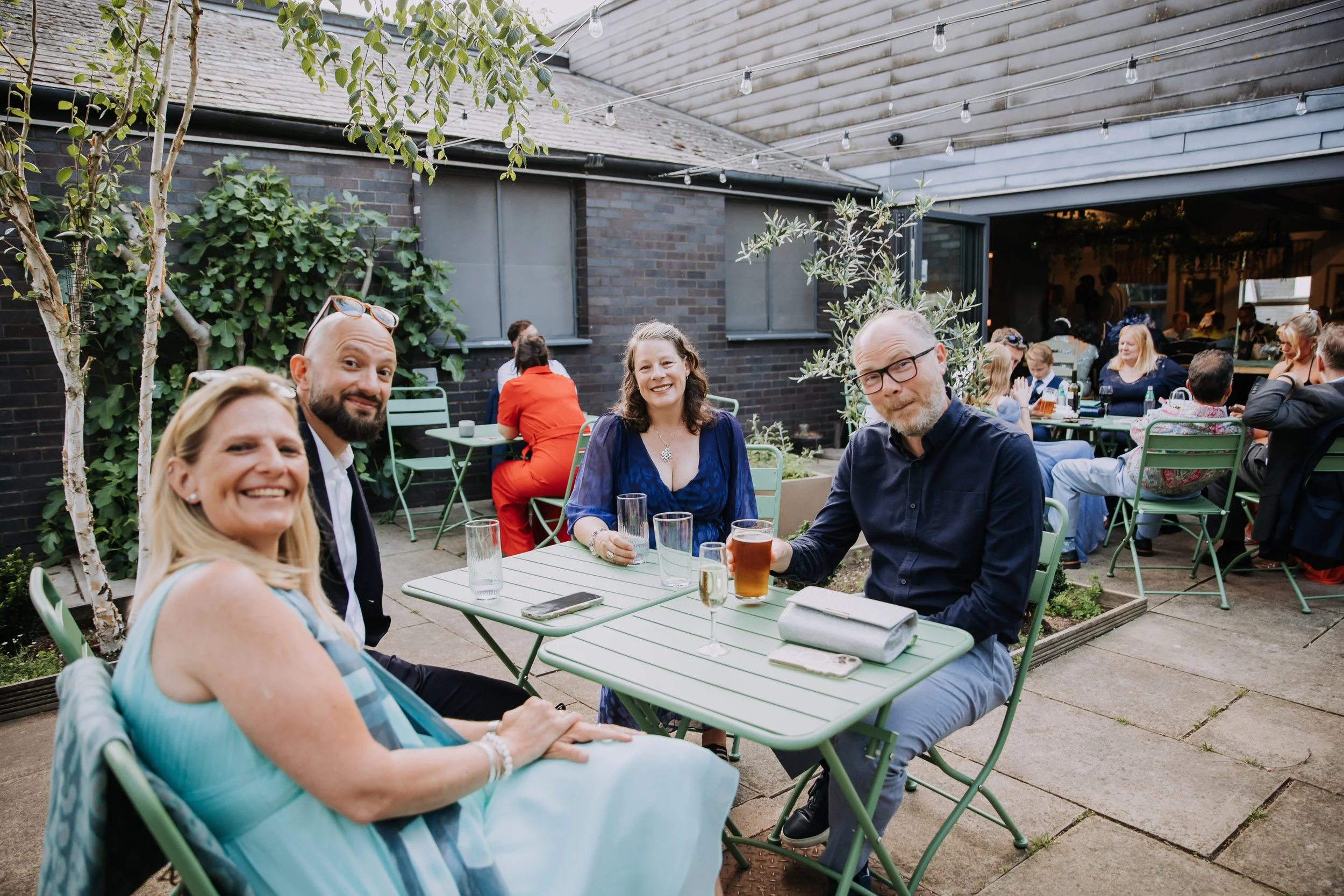 Four people sitting at a table outdoors at a social gathering, smiling and holding drinks, with more guests in the background, in a patio setting.