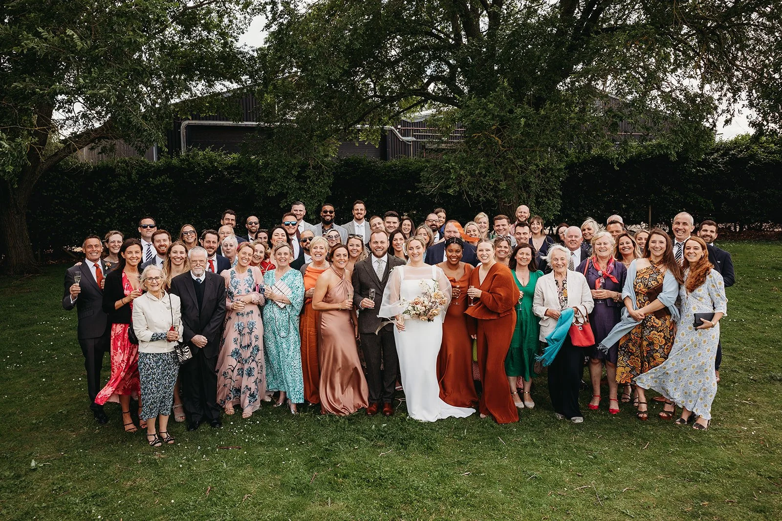 A large group of people gathered outdoors for a wedding celebration on a grassy area with trees in the background. The bride and groom are standing at the center, smiling, with the bride holding a bouquet.