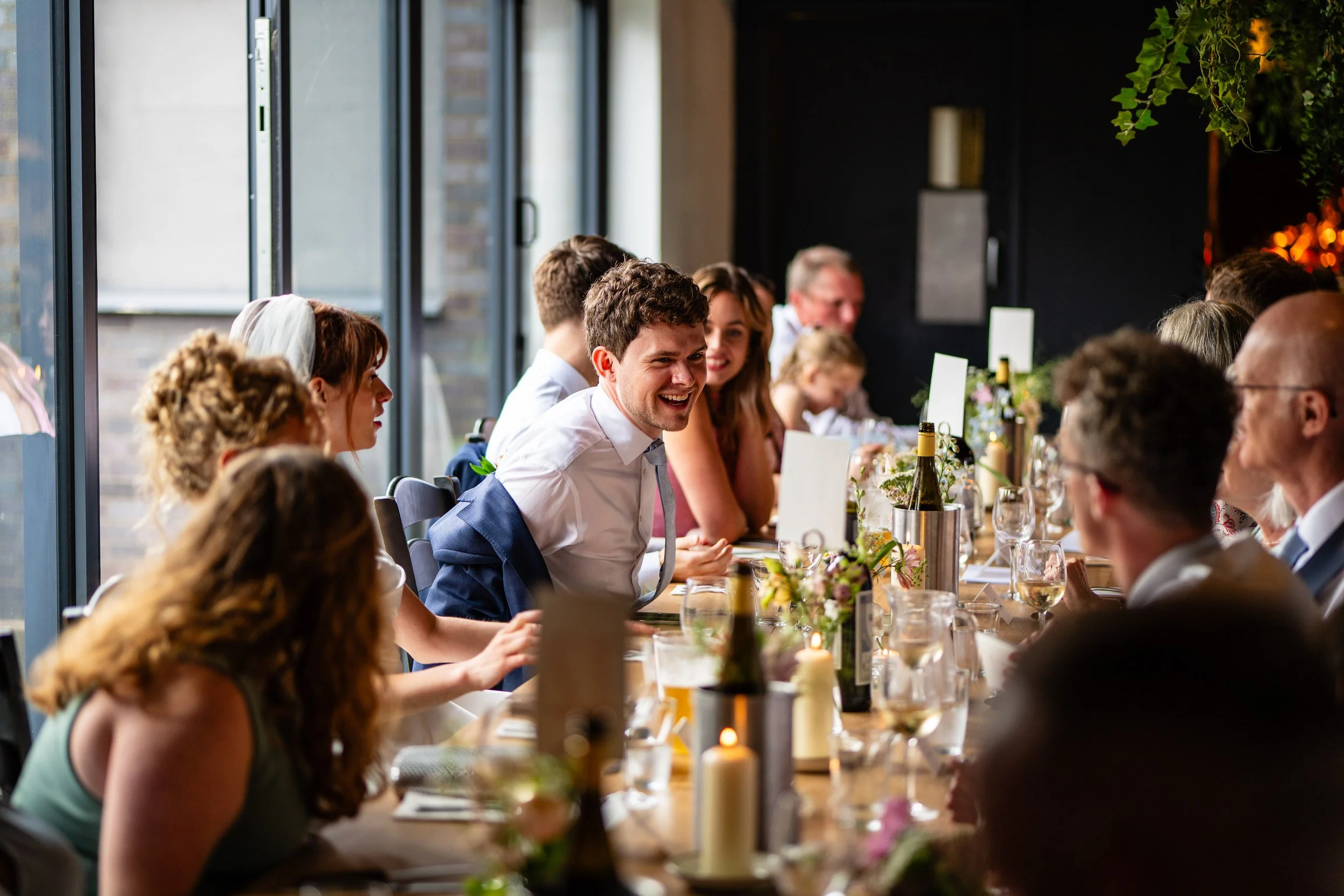 People sitting at a long dining table during a celebration, smiling and engaging with each other, with bottles of wine and floral centerpieces on the table inside a modern, well-lit venue.