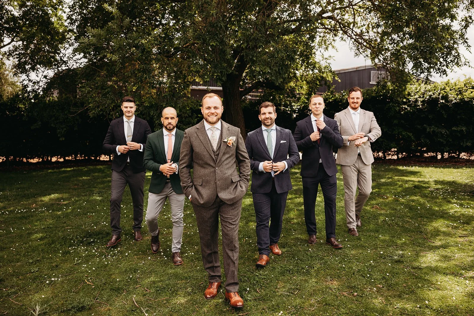 A group of seven men dressed in suits standing outdoors on grass in front of a large tree, smiling for a photo.