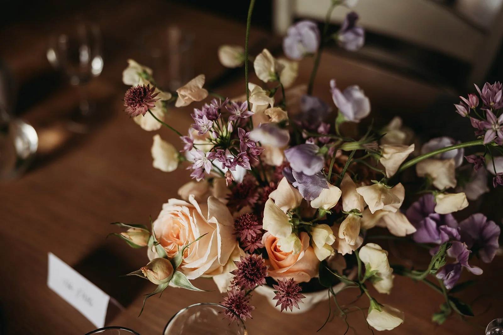 A floral arrangement with peach roses, purple and cream flowers, on a wooden table.