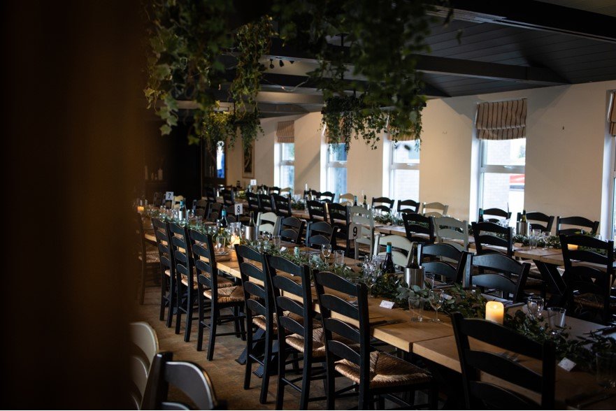 Long dining table set for a formal event with wine bottles, glasses, and greenery decoration, in a well-lit room with multiple windows and hanging plants.