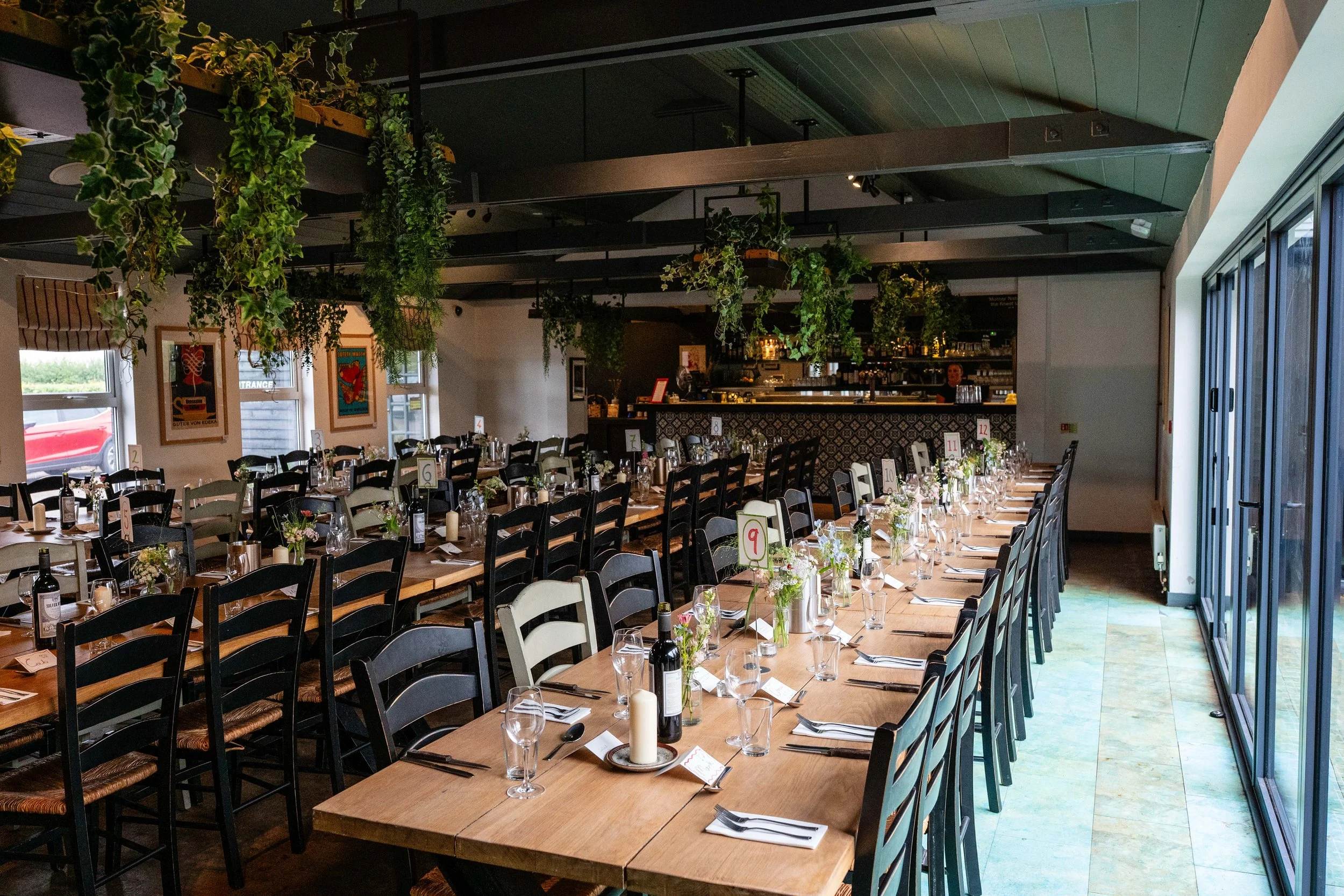 Interior of a restaurant set up for a large event with long tables, wine bottles, glasses, candles, flowers, and numbered table markers, with hanging plants and artwork on the walls.