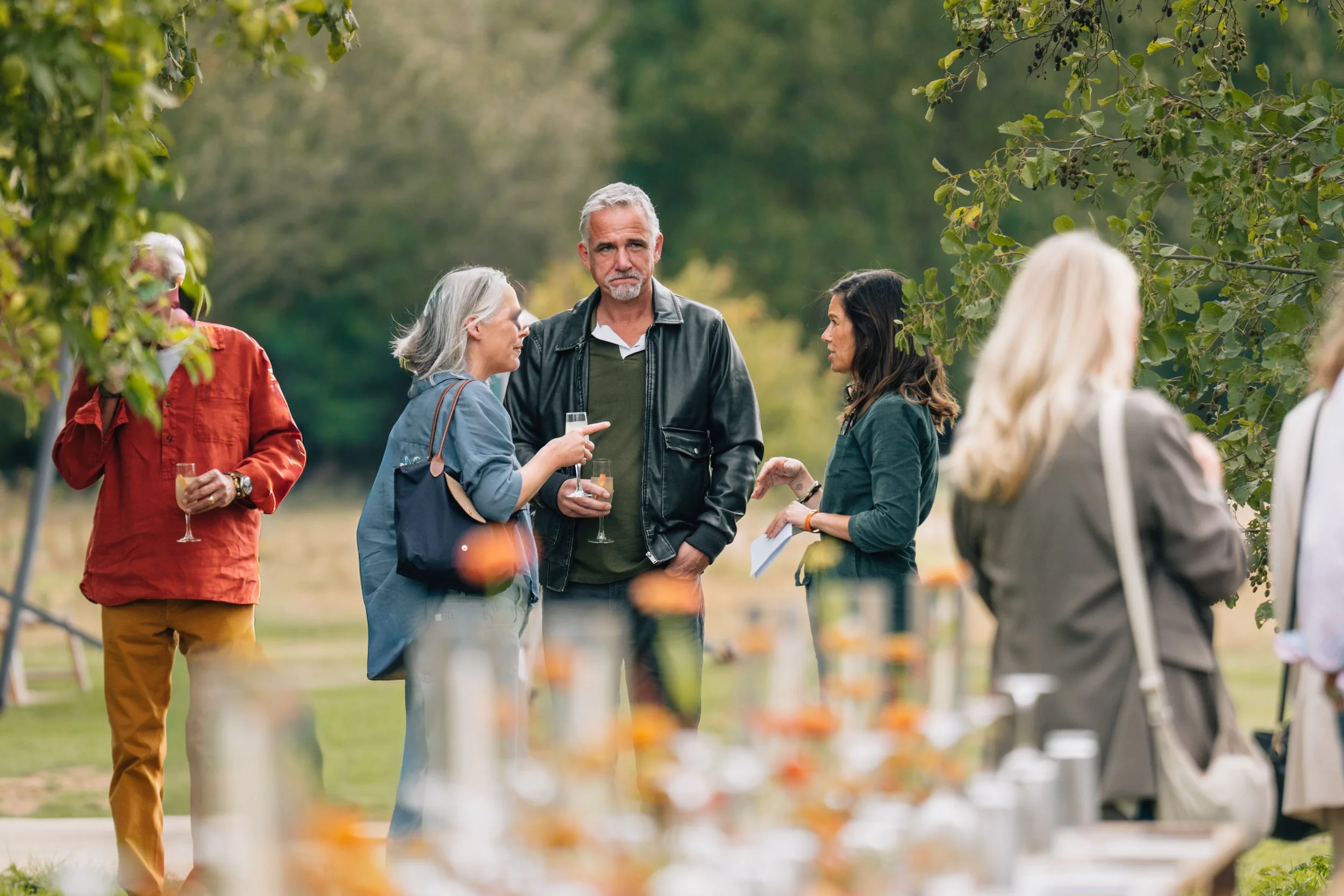 People gathered outdoors at a social event, some holding glasses of wine, engaged in conversation in a park-like setting with trees and blurred foreground objects.