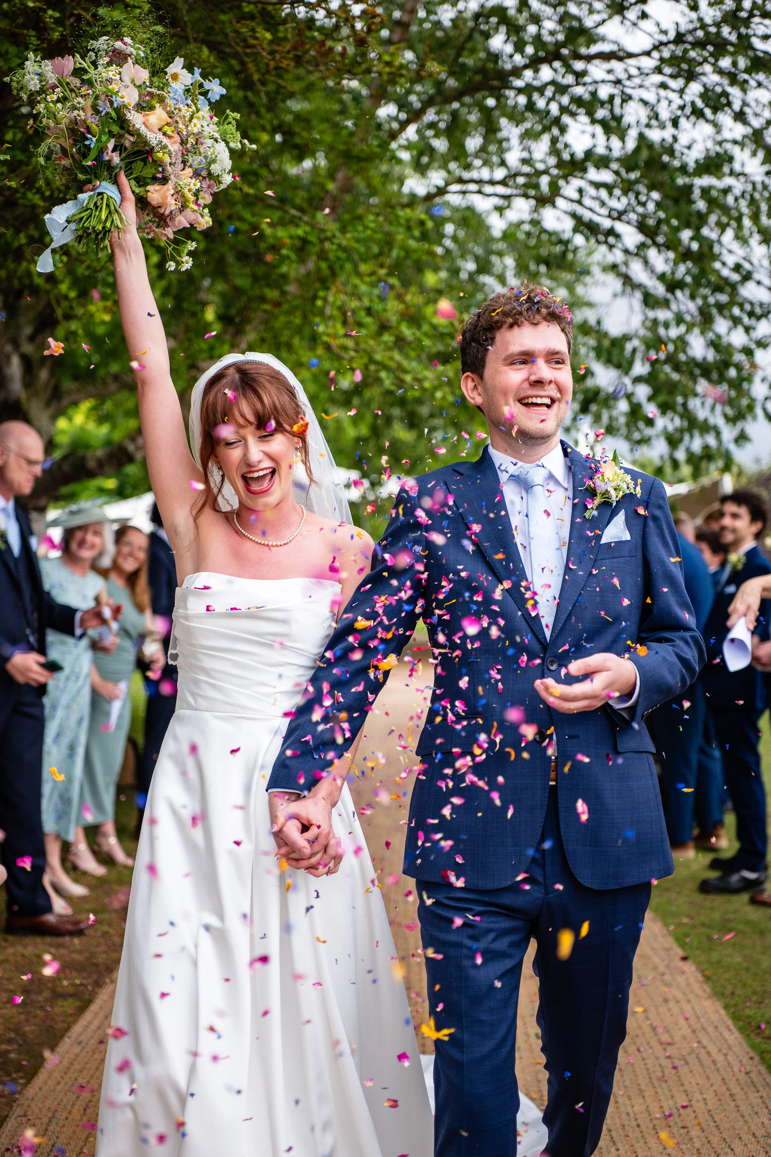 A newlywed couple walking hand in hand outdoors surrounded by friends and family, throwing colorful confetti during their wedding celebration, with trees and guests in the background.
