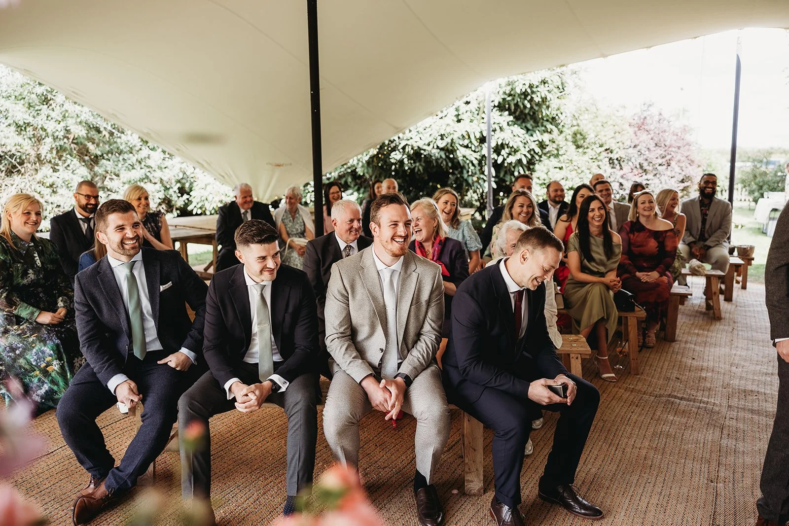 Group of people seated under a large outdoor tent, dressed in formal attire, smiling and laughing during a wedding ceremony.