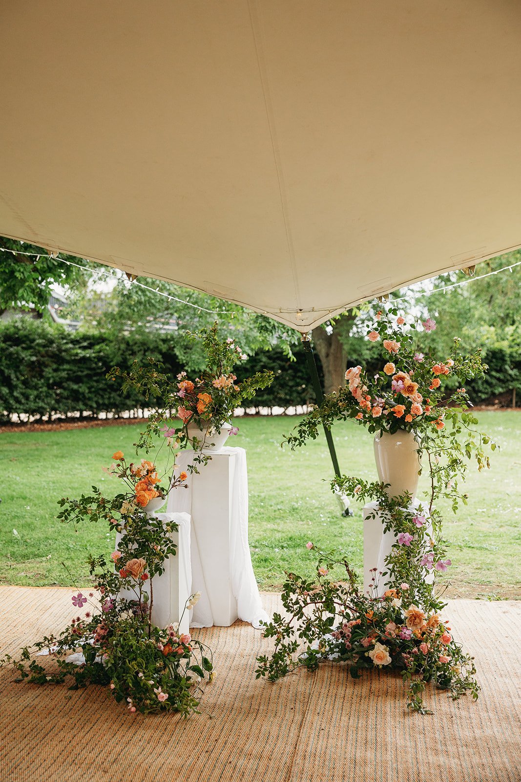 Decorative floral arrangements with pink, orange, and white flowers and greenery on white pedestals under a beige canopy in a garden setting.