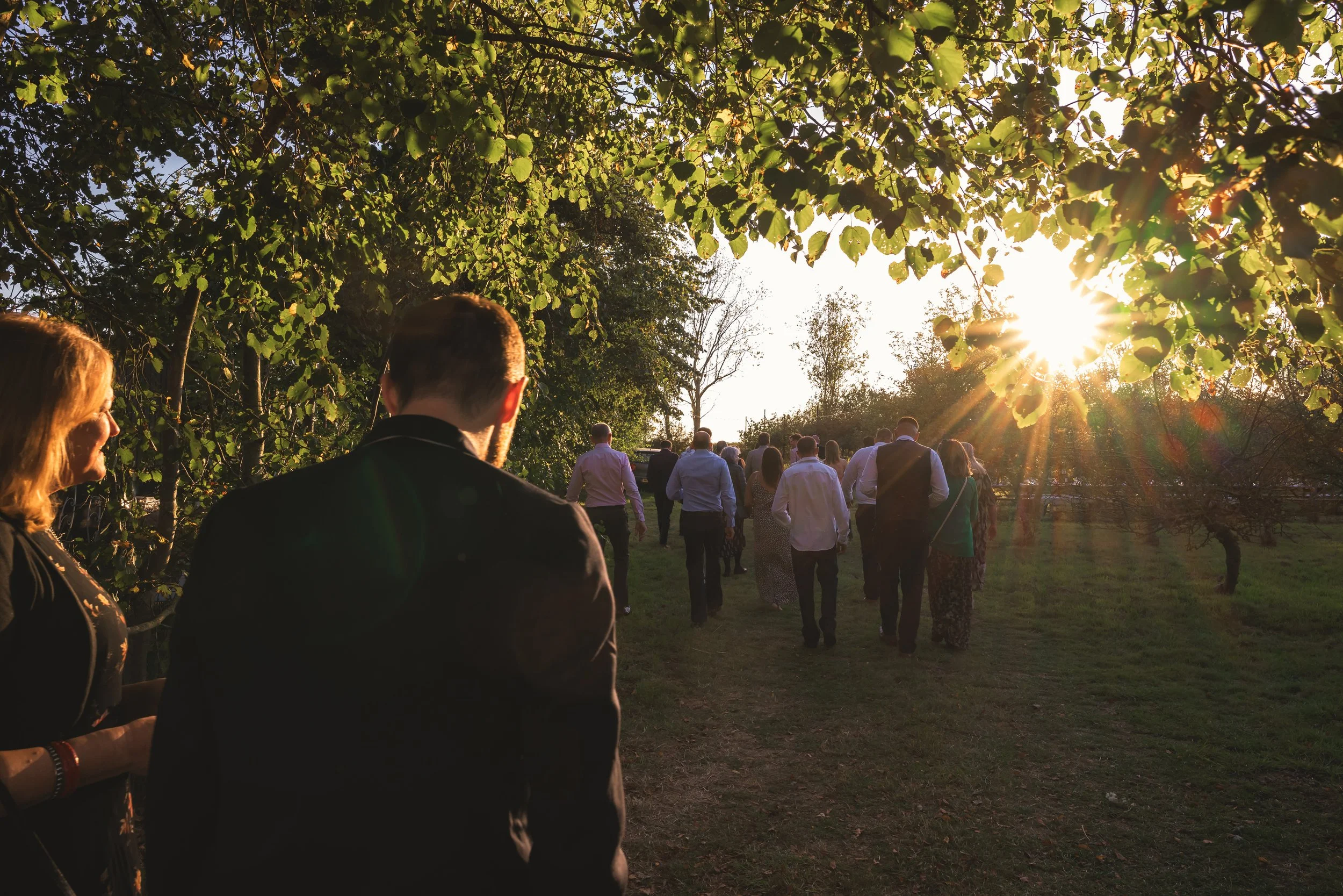 A group of people walking through a green outdoor area during sunset, with trees and sunlight in the background, some of whom are dressed formally.