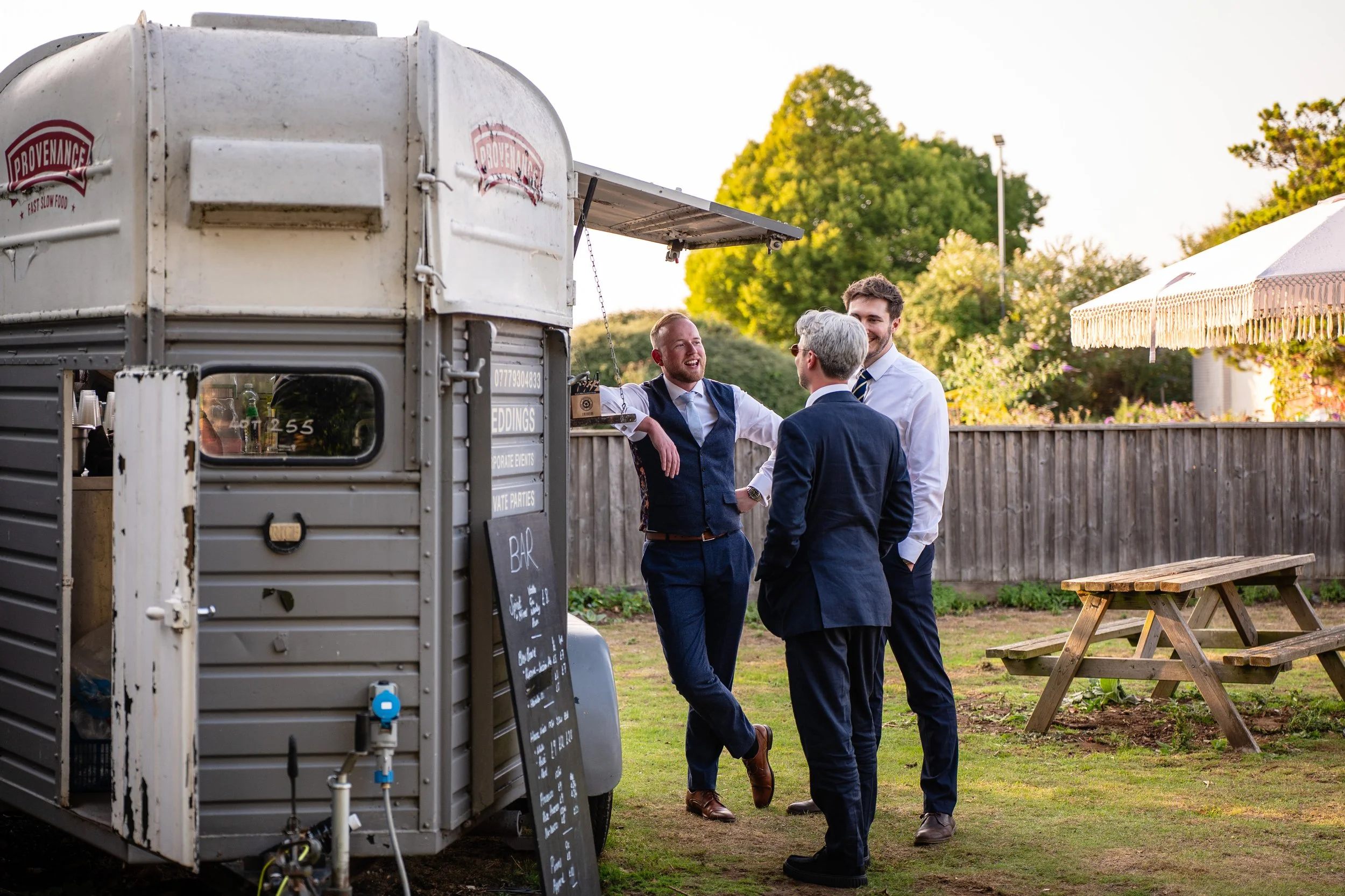 Three men in formal attire talking and laughing next to a vintage mobile bar at an outdoor event during sunset, with trees, a fence, and a picnic table in the background.