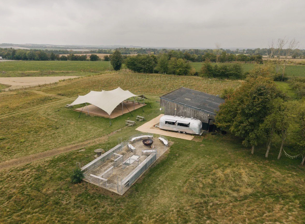 Aerial view of a rural outdoor area with a large white canopy, a weathered barn, a vintage silver camper, a fenced animal enclosure, and large trees, with expansive fields and overcast sky in the background.