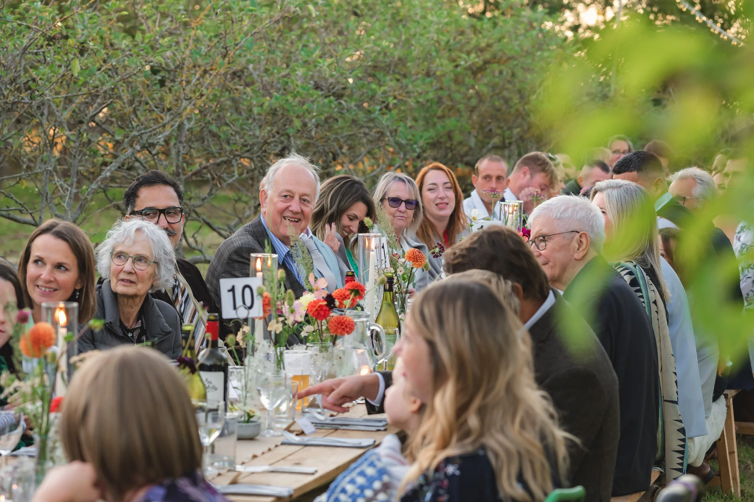 People gathered at a long outdoor dining table decorated with flowers and candles, smiling and engaging in conversation during a daytime event in a garden.