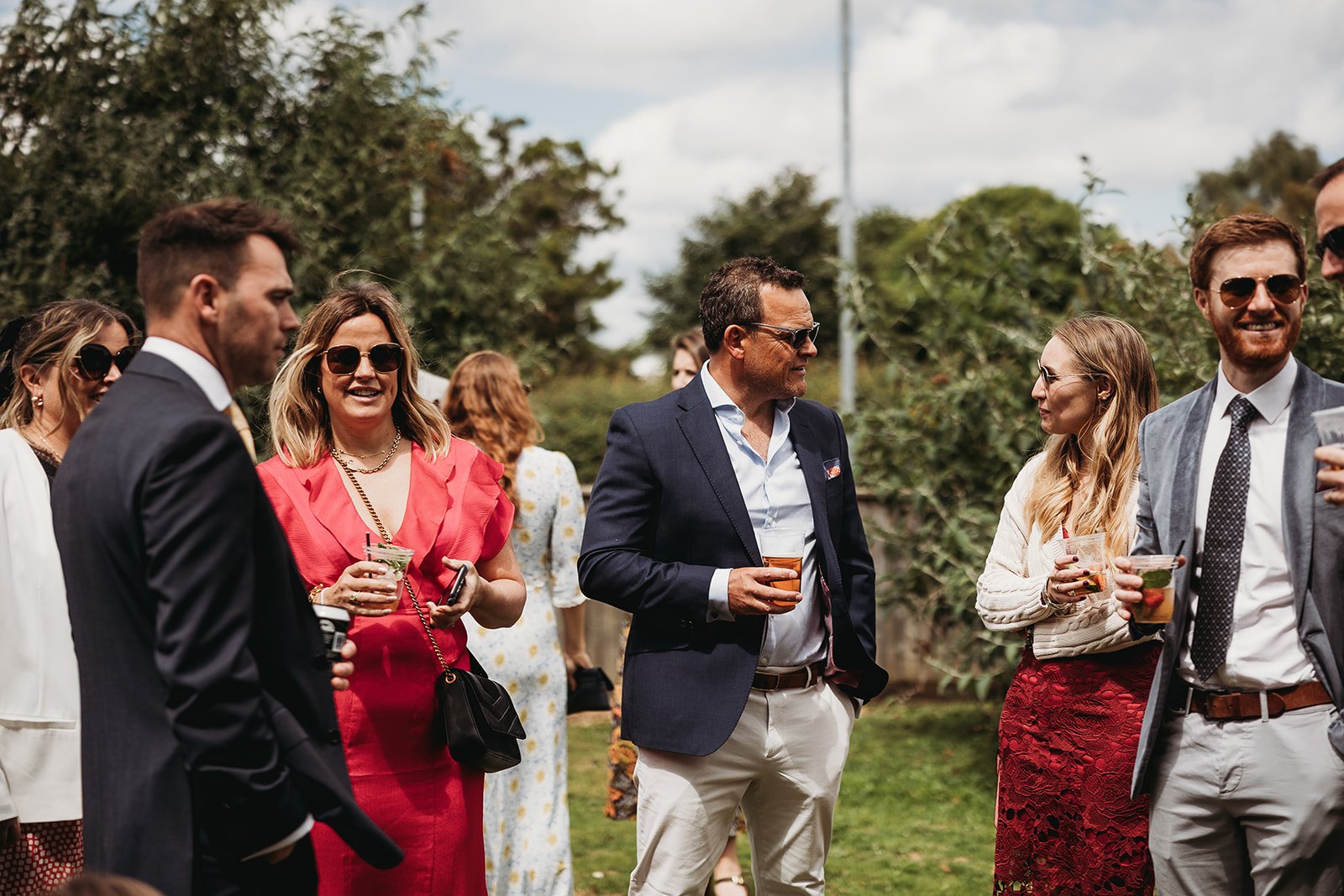 Group of well-dressed people socializing outdoors, some holding drinks, in a garden or park setting on a partly cloudy day.