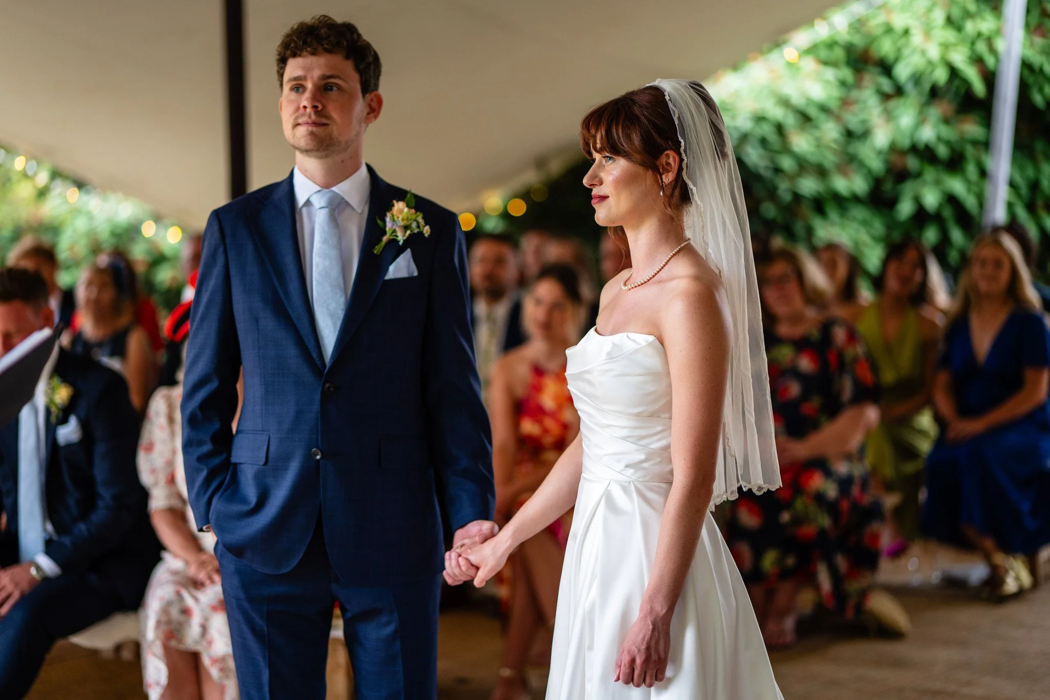 Bride and groom holding hands during wedding ceremony with guests seated in the background under a tent.