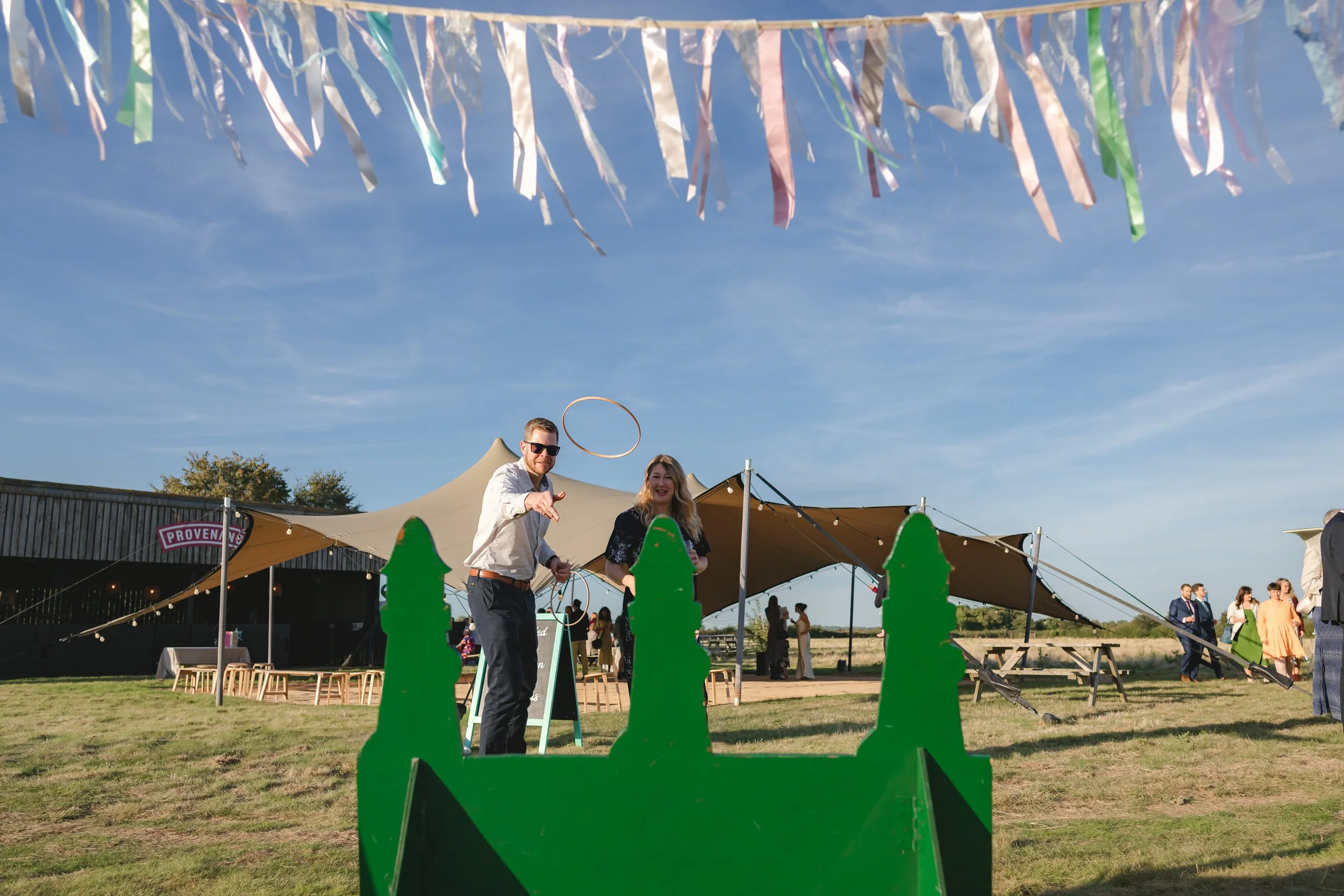 People playing ring toss game at outdoor event with a large tent, string lights, and colorful banners overhead.