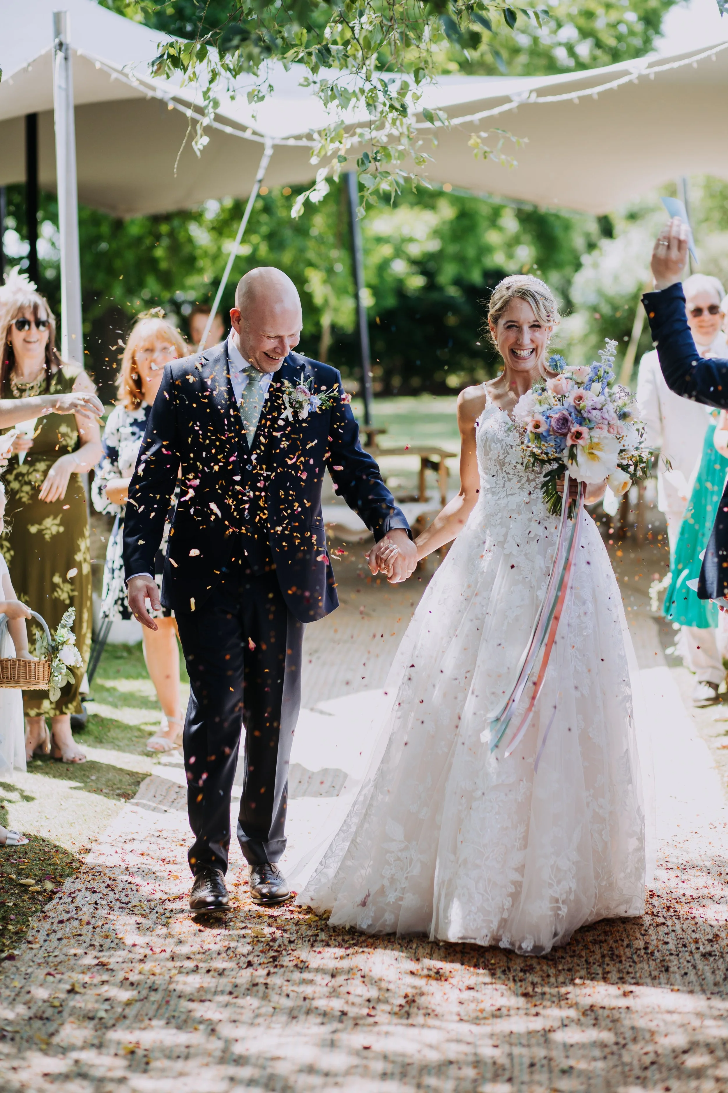 A newlywed couple walking hand-in-hand, smiling, after their wedding ceremony with guests celebrating and throwing confetti outdoors on a sunny day.