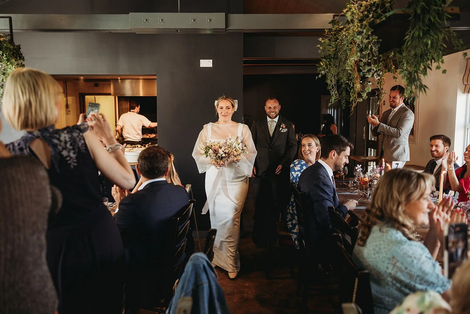 Bride walking into a wedding reception with her groom, surrounded by seated guests clapping and taking photos, in a decorated indoor venue.