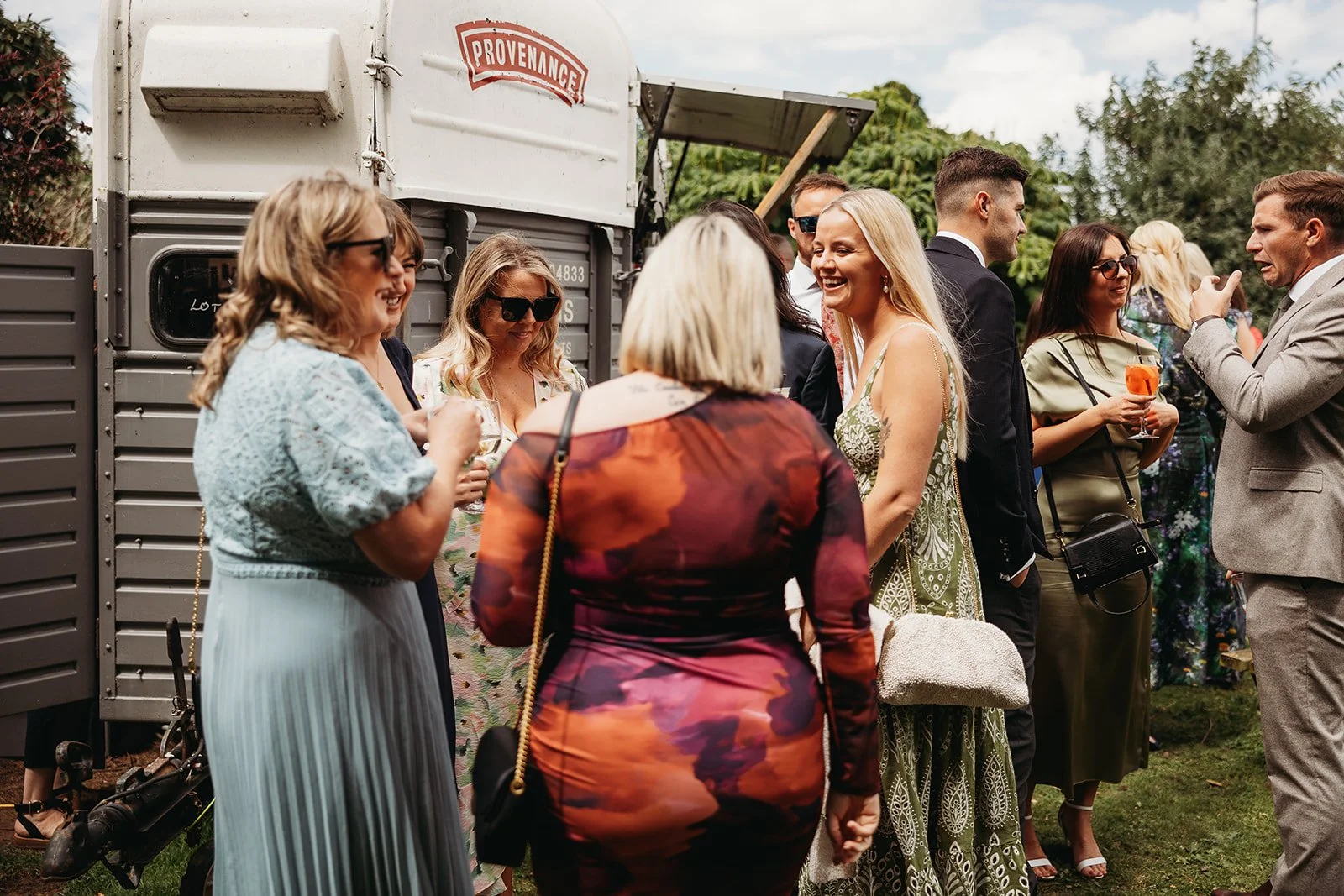 Group of people socializing outdoors at a festive event, with some holding drinks and talking cheerfully, near a vintage trailer.