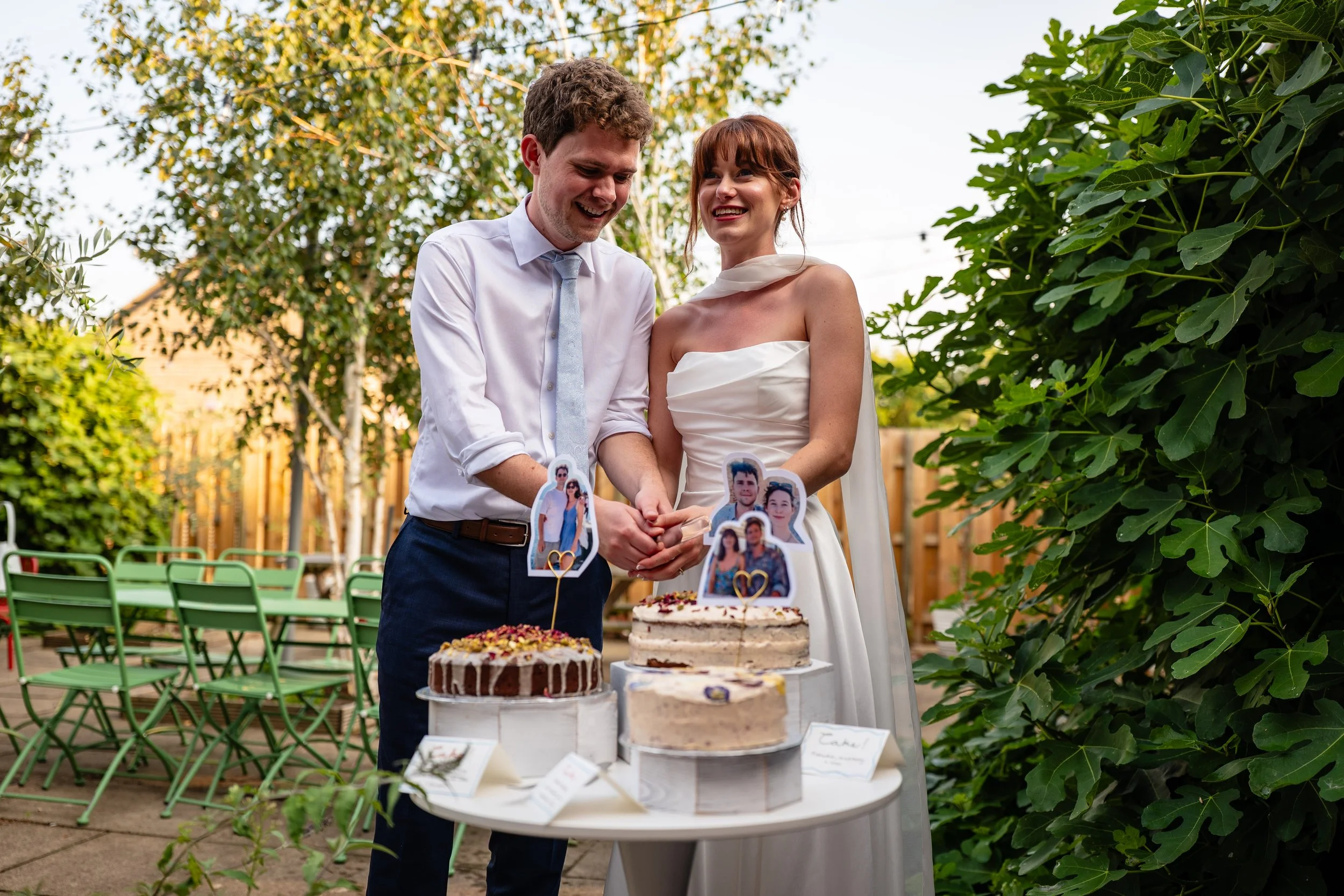 A newlywed couple cutting their wedding cake outdoors, surrounded by trees and green chairs, with photo toppers on the cakes.