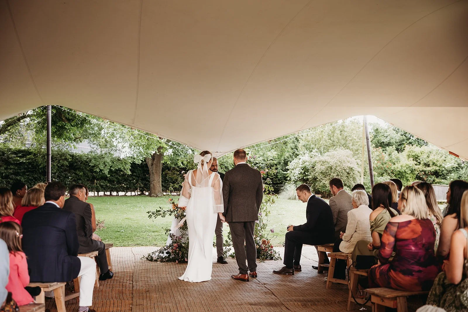 A couple getting married outdoors under a large canopy, surrounded by seated guests, with trees and greenery in the background.