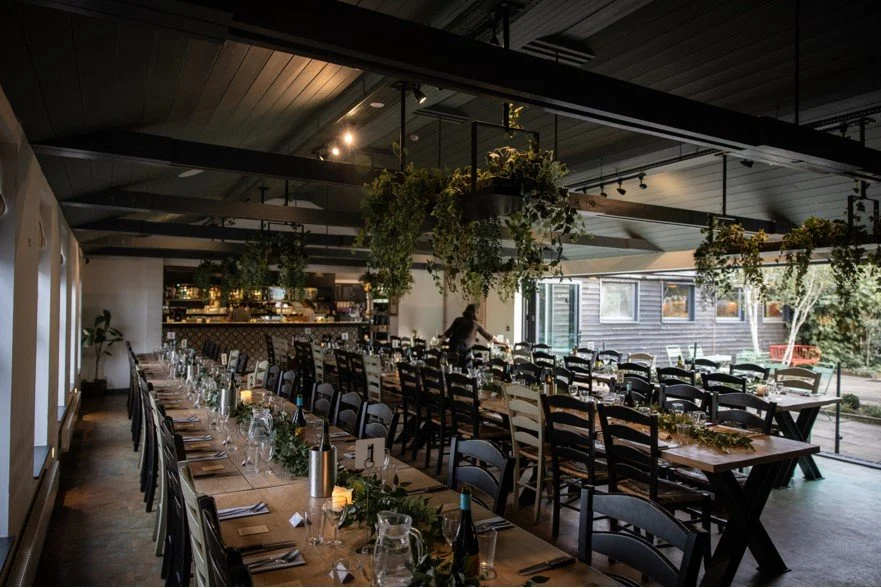 Dining area with long, decorated tables set with plates, glasses, and cutlery, surrounded by black chairs, with hanging plants overhead and large windows letting in natural light.